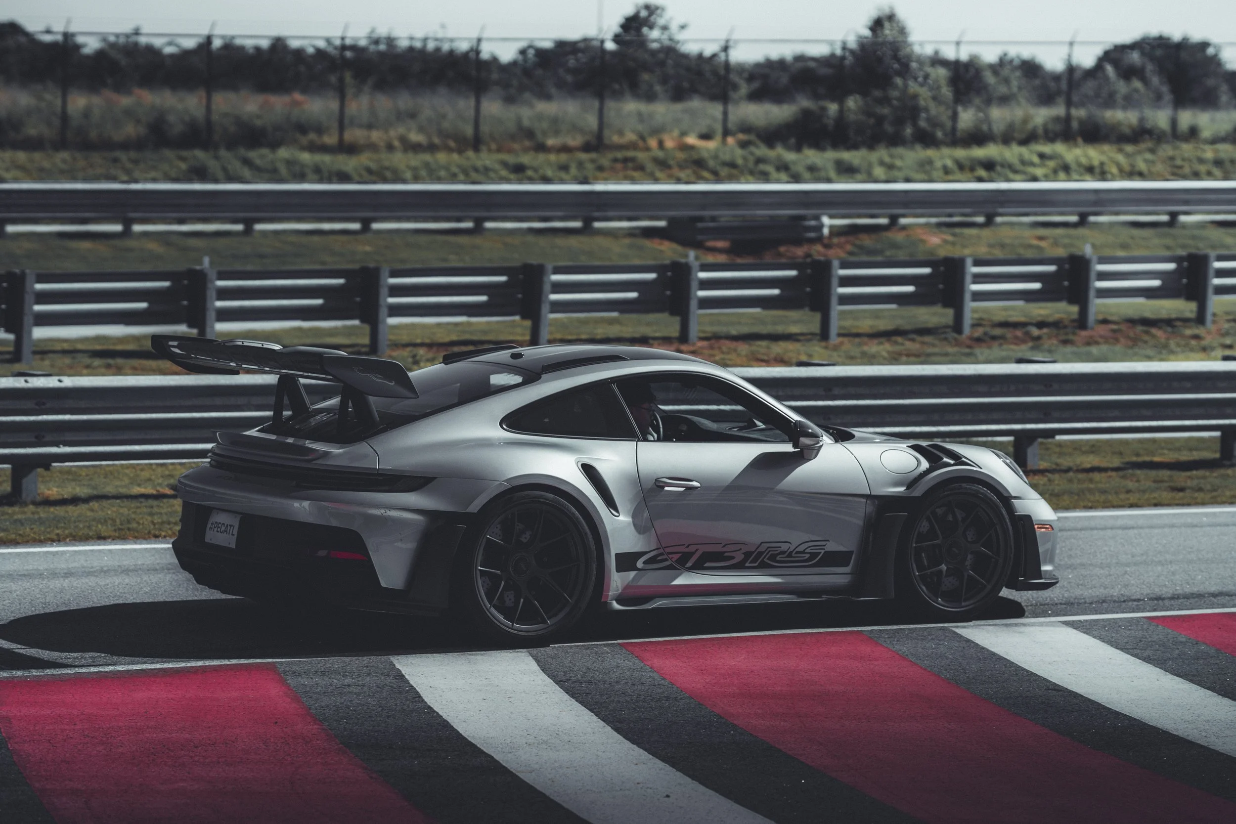 A silver sports car with a large rear wing, racing decals, and black wheels driving on a race track with red and white curbs, with a grassy area and trees in the background.