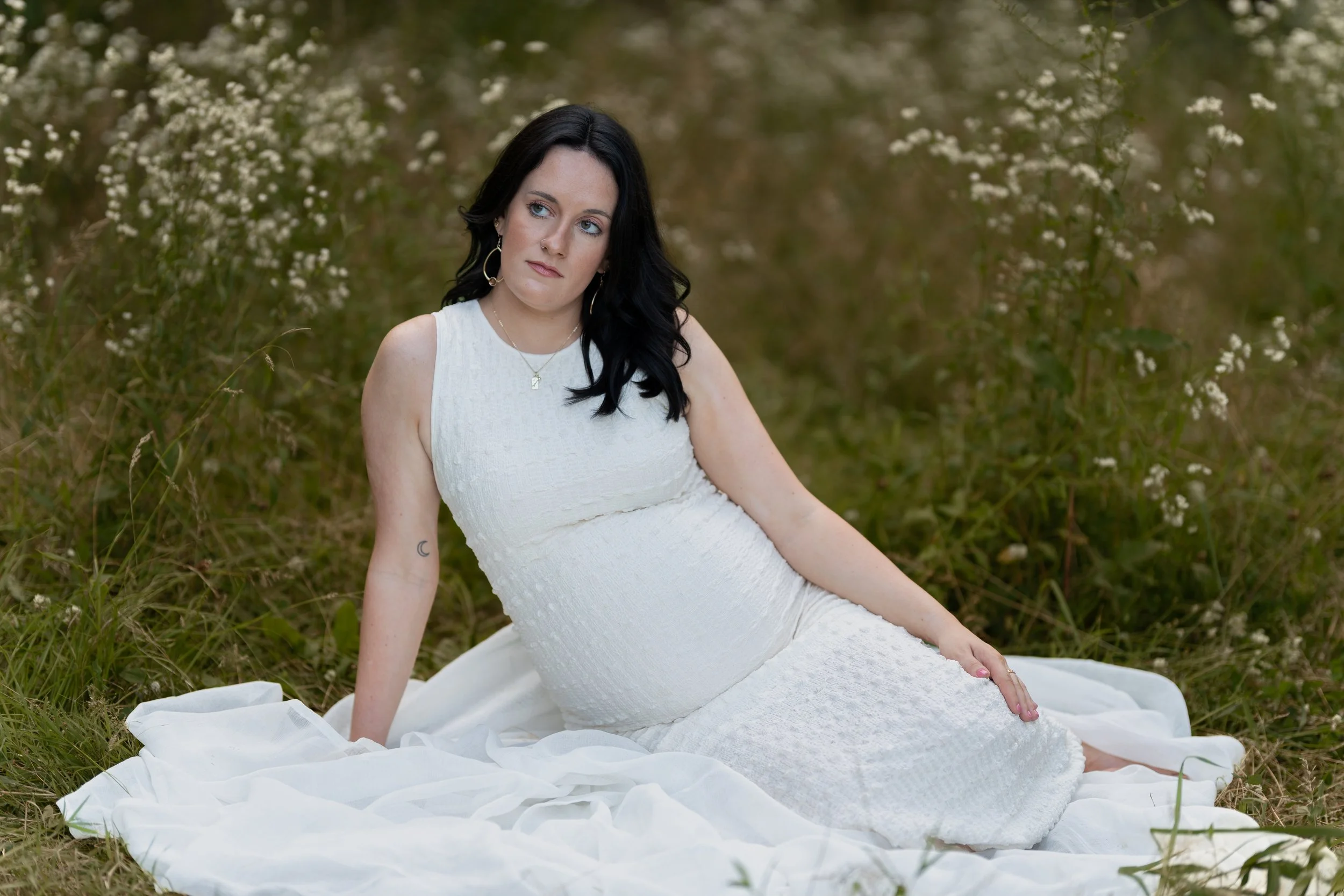 A woman with black hair and light skin, wearing a white dress, sitting on the grass in a field of small white flowers, with a contemplative expression.