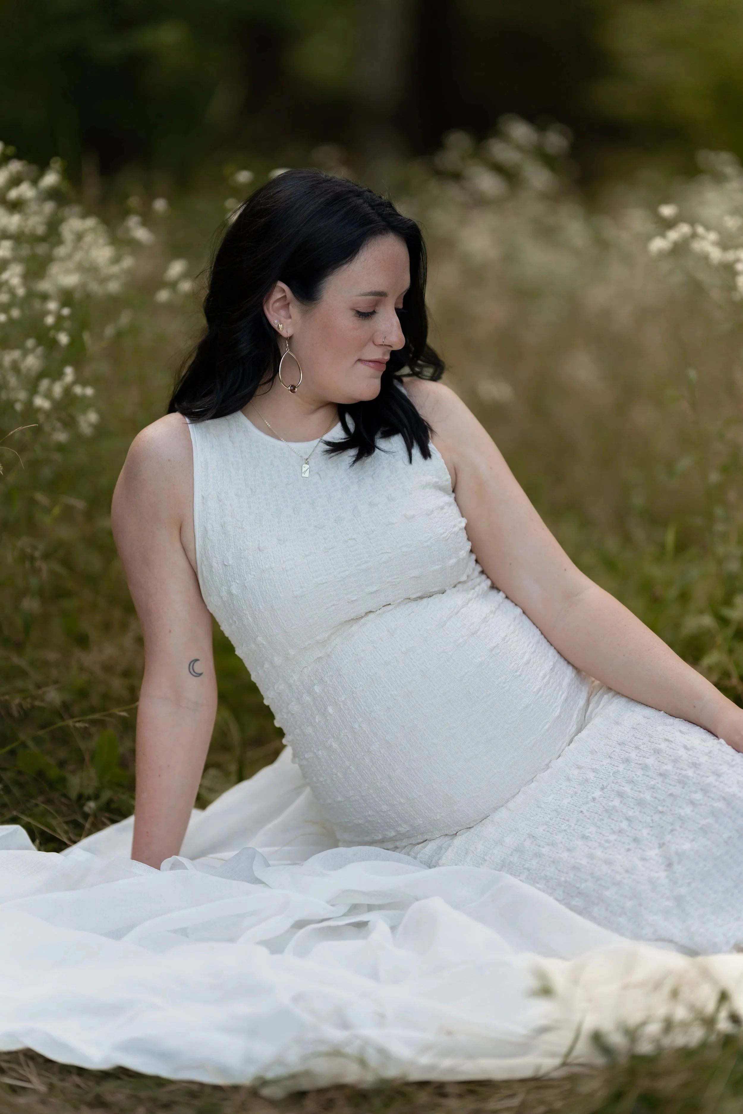 Woman with black hair, wearing a white dress, sitting outdoors on grass with white flowers in the background, looking down with a serene expression.