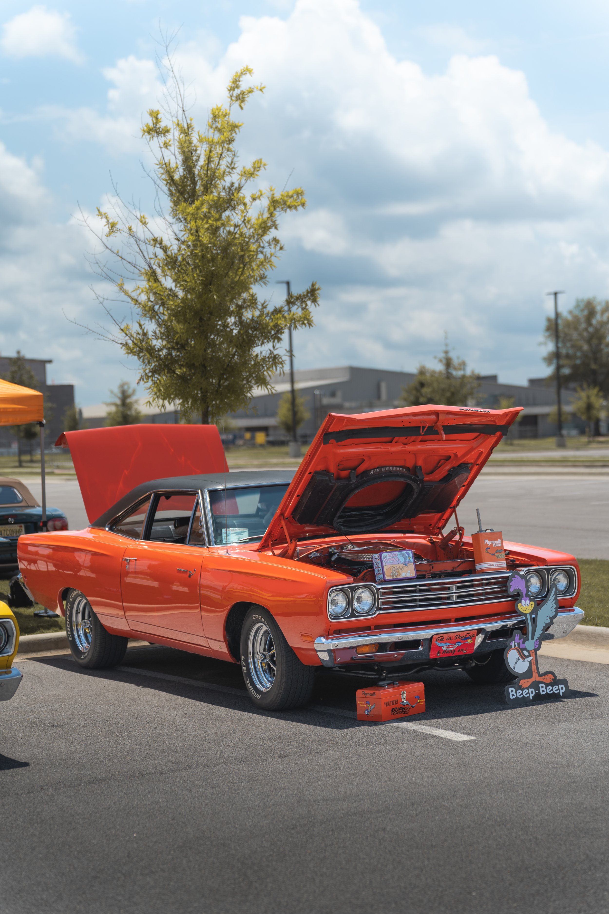 Orange vintage car with its hood open on display at an outdoor event, decorated with cartoon character cutouts and a sign that says "Get in, Shut up, and Hang On."
