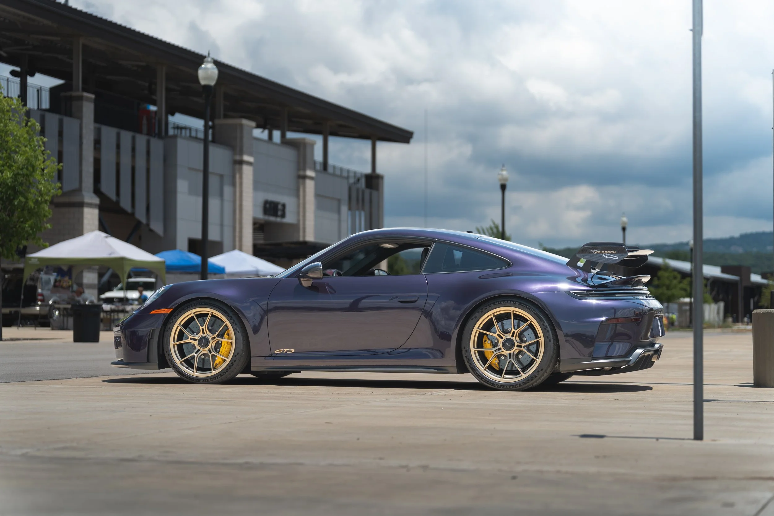 A dark purple Porsche 911 GT3 parked outdoors under cloudy skies, with gold rims and a spoiler, near tents at a public event.