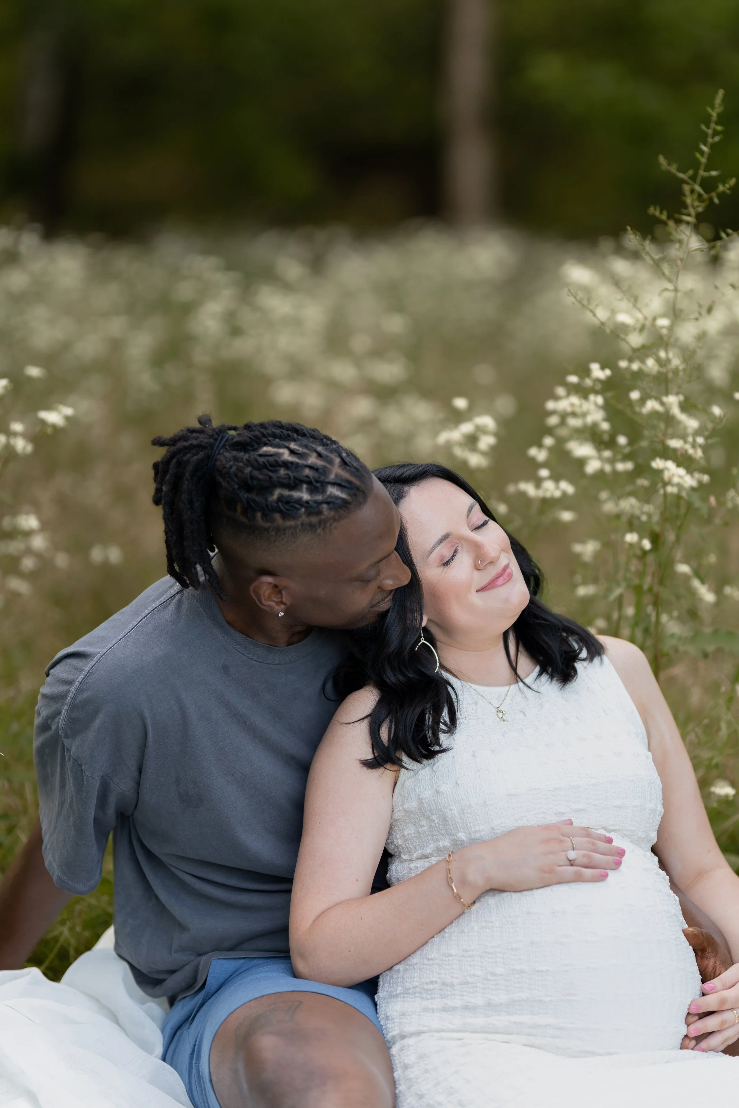 A couple sitting outdoors in a field of white flowers, with the man kissing the woman on the forehead as she smiles peacefully, her hand resting on her pregnant belly.