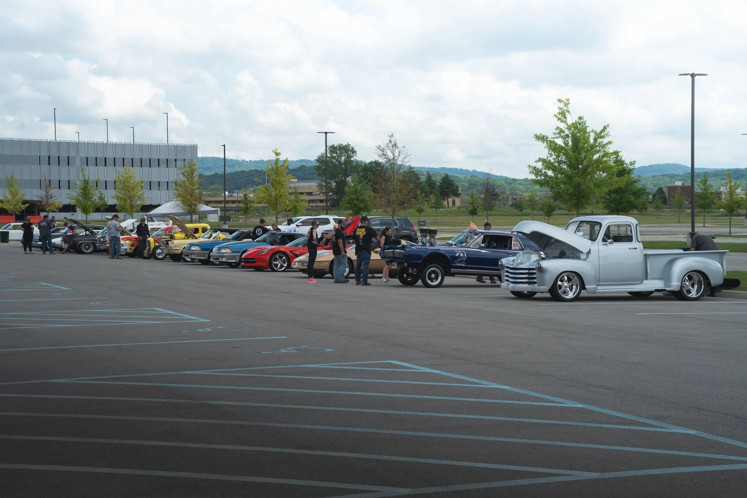 A parking lot with a line of vintage and sports cars on display, with people walking around and looking at the vehicles, and a large modern building and hills in the background.