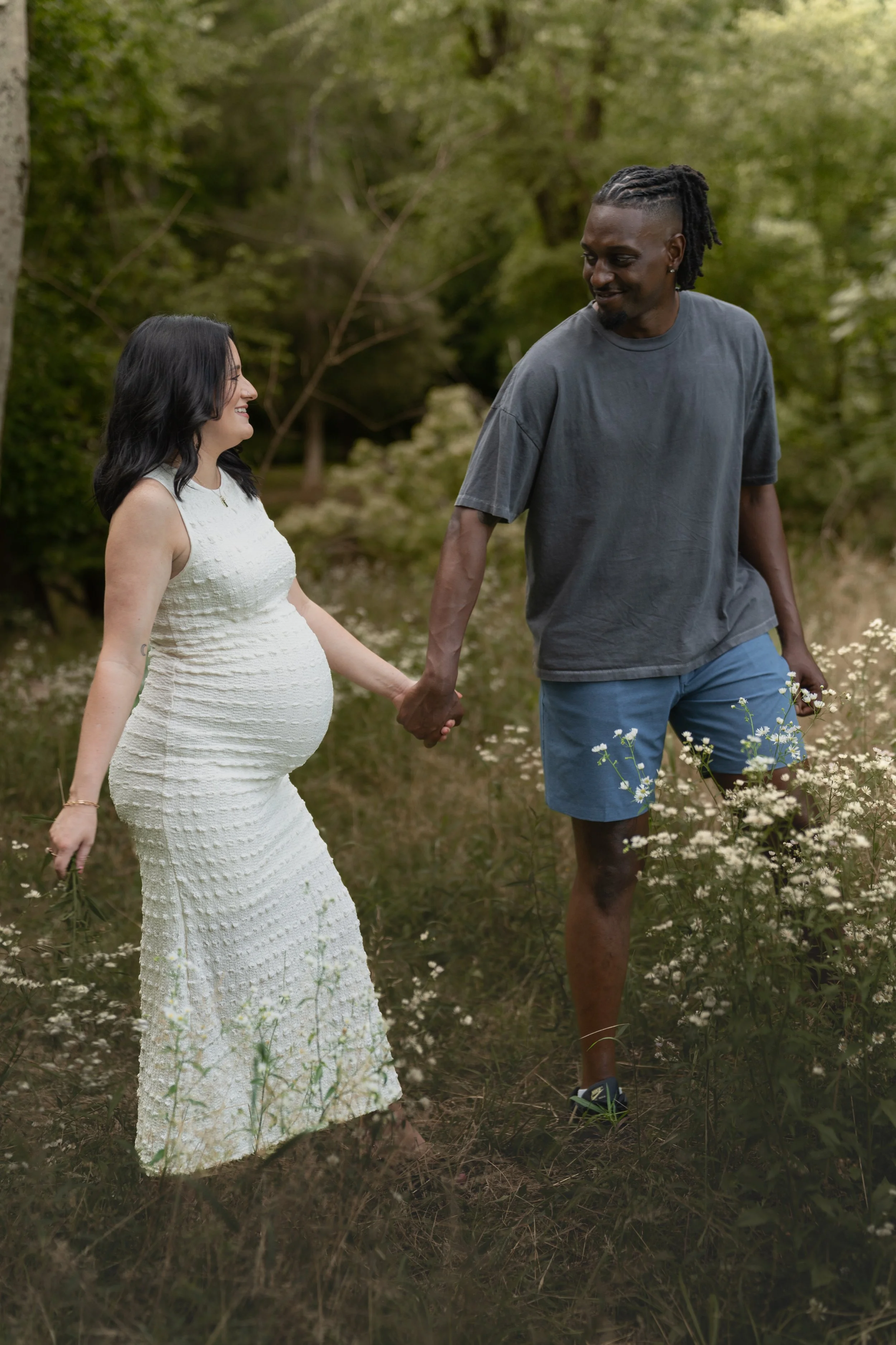 A pregnant woman and a man holding hands while walking in a field of wildflowers in a wooded area, smiling at each other.
