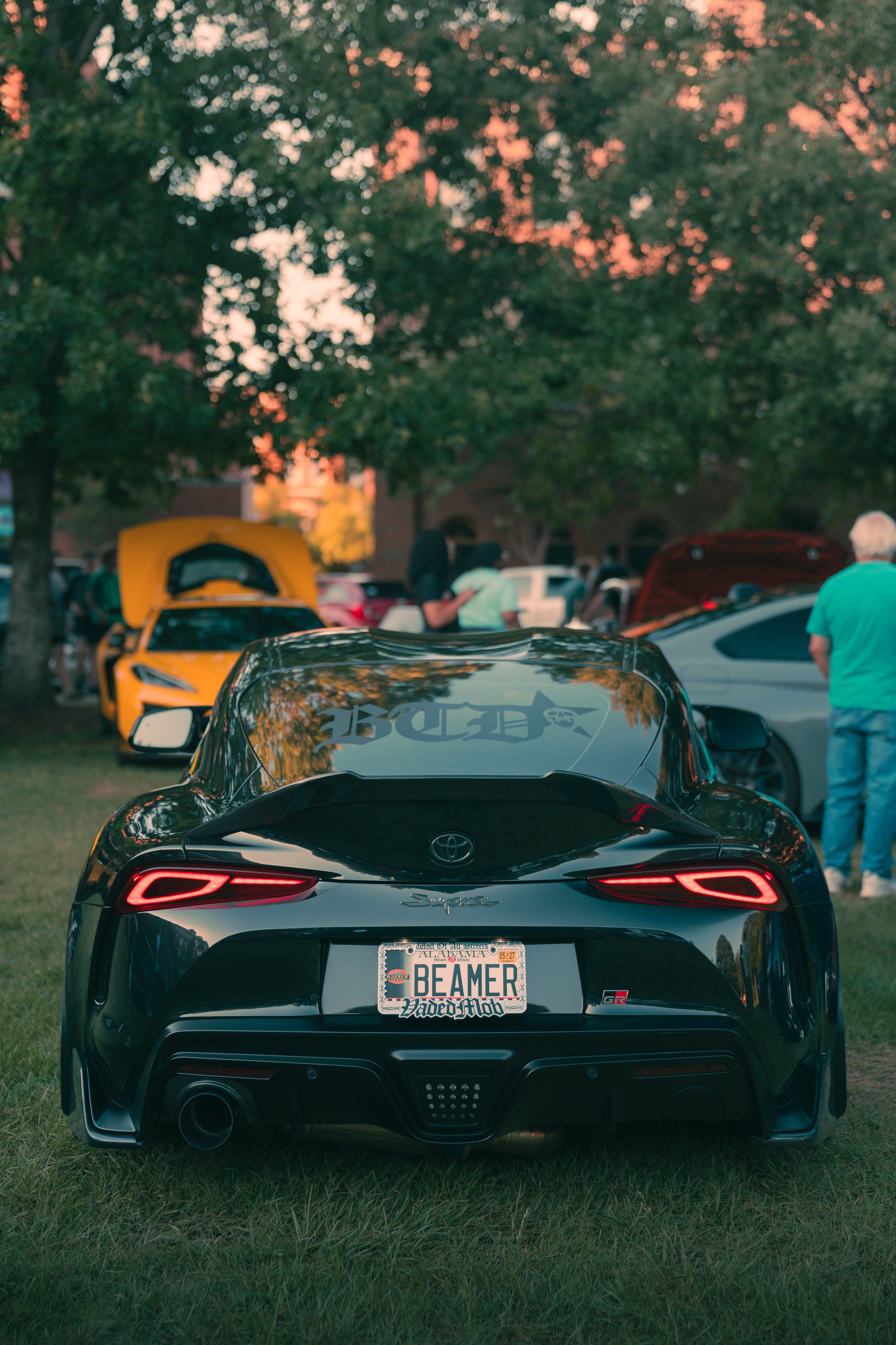 Rear view of a black Toyota Supra sports car with a personalized license plate that reads 'BEAMER', parked on grass at a car meet. The background shows other cars, open hoods, and a group of people under a large tree at sunset.