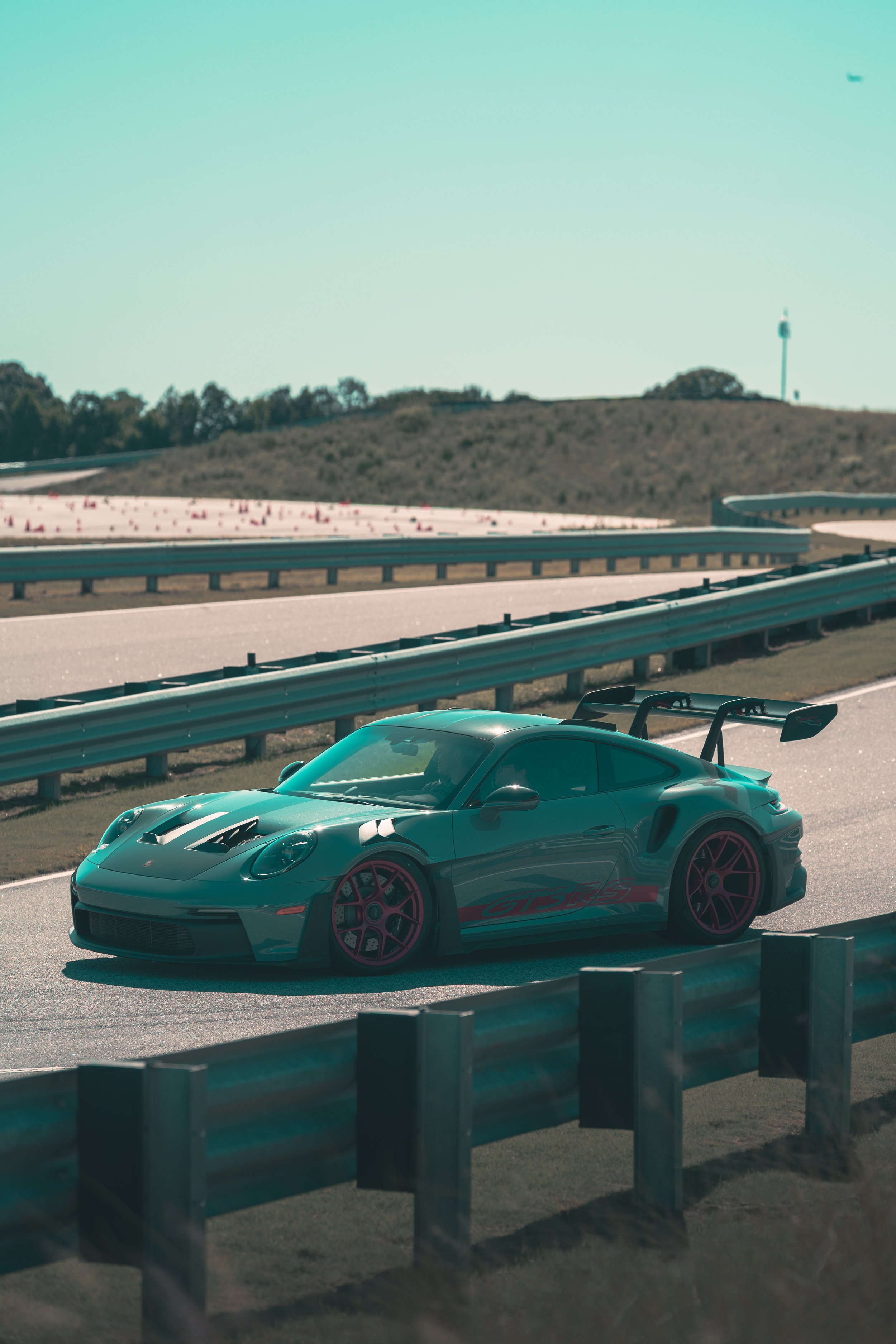 A green sports car with red accents parked on a road near barriers, with a hill and wind turbine in the background under a clear sky.