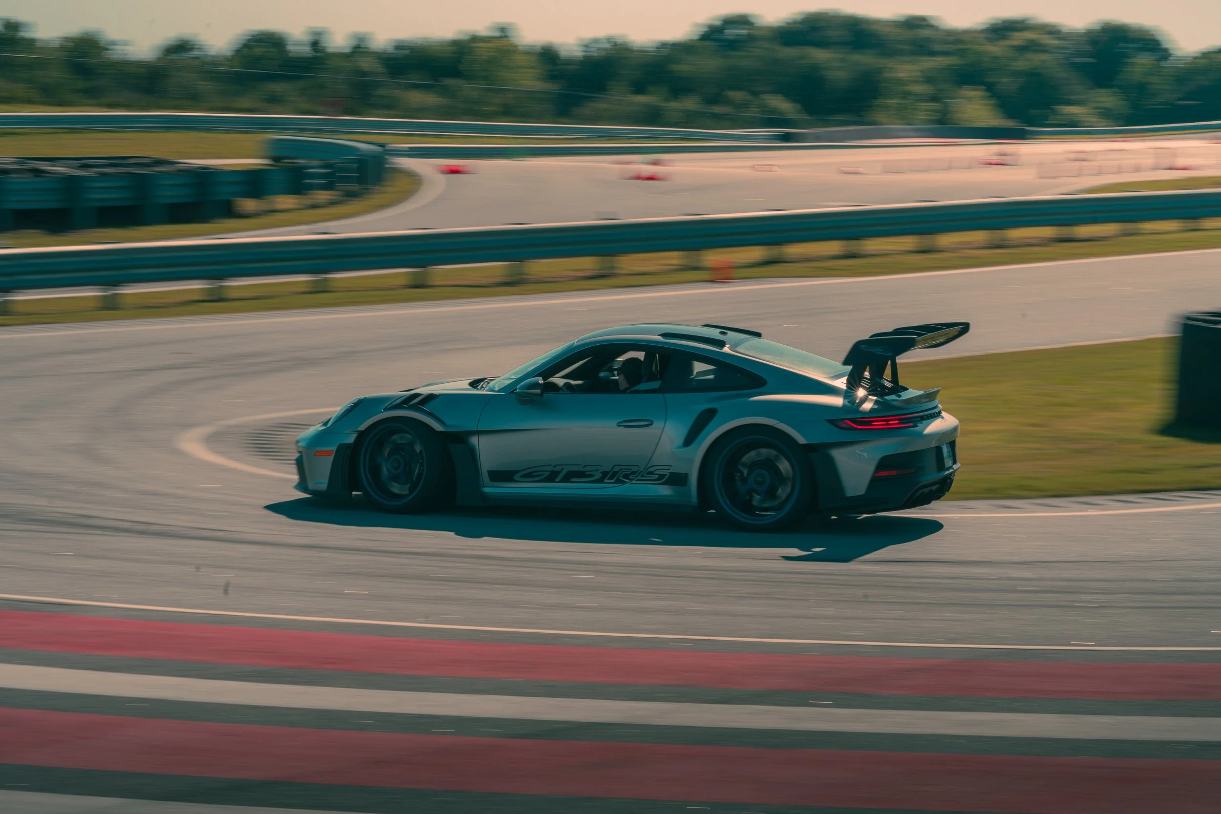 A silver Porsche 911 GT3 RS race car on a racetrack, taking a turn with tire barriers and guardrails in the background, and trees in the distance.