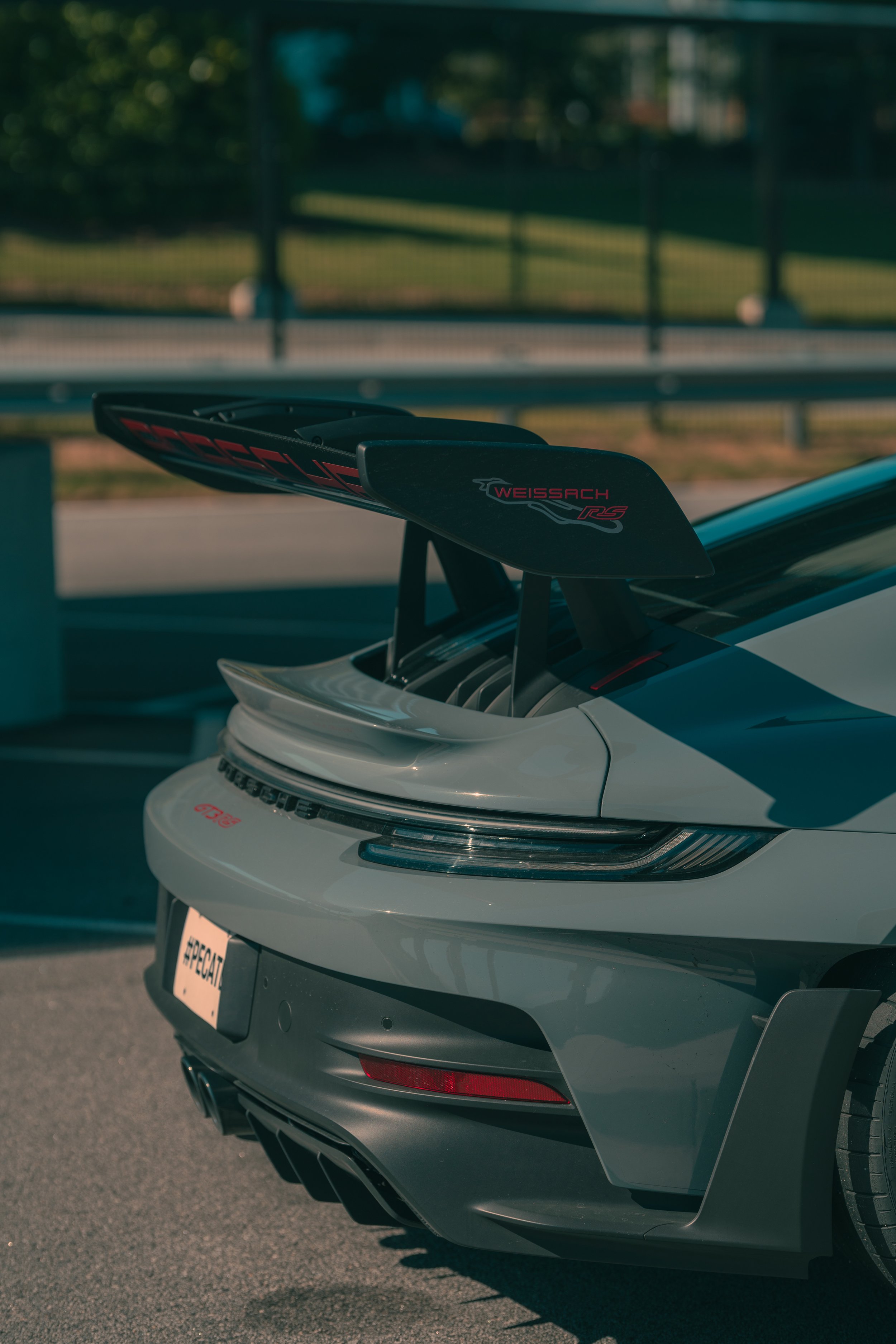 Close-up of the rear end of a white sports car with a large black and red rear wing spoiler, parked on a street with a metal fence and trees in the background.