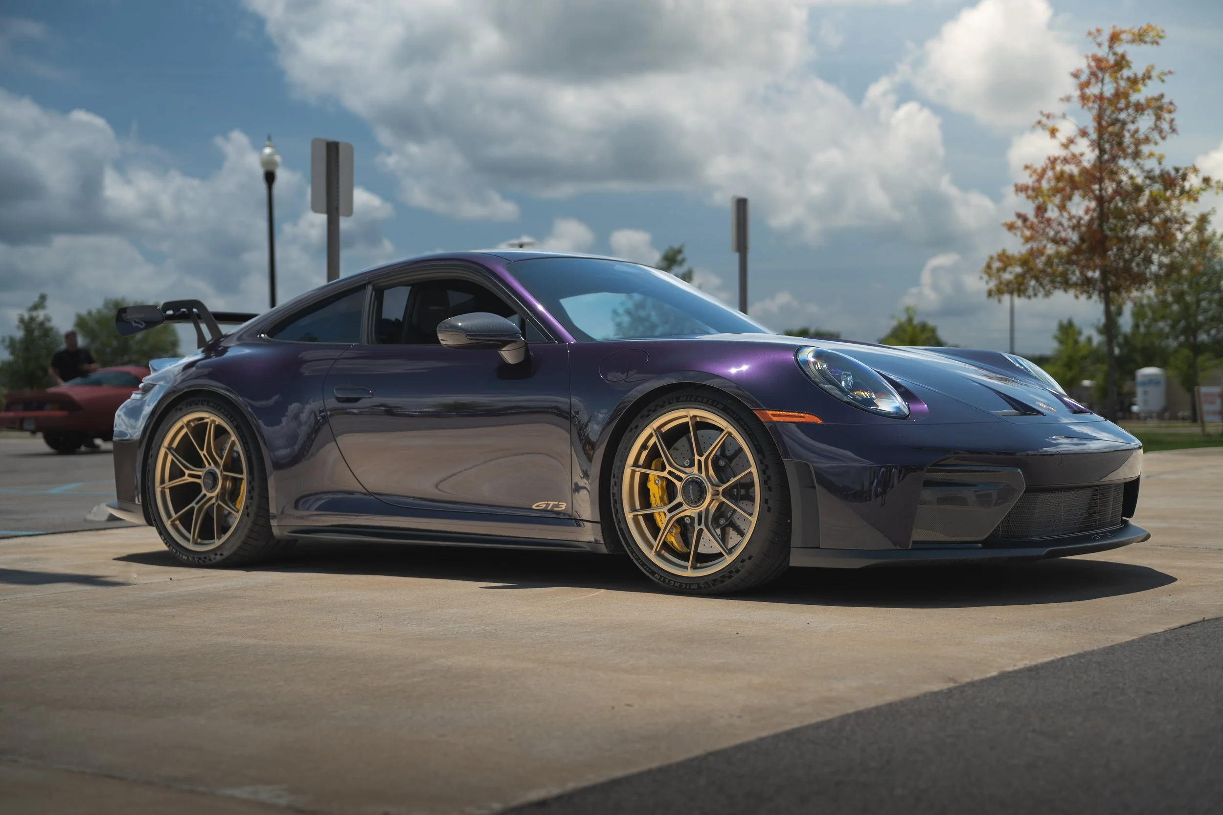 A dark purple Porsche 911 GT3 sports car parked outdoors on a sunny day, with some trees and a partly cloudy sky in the background.