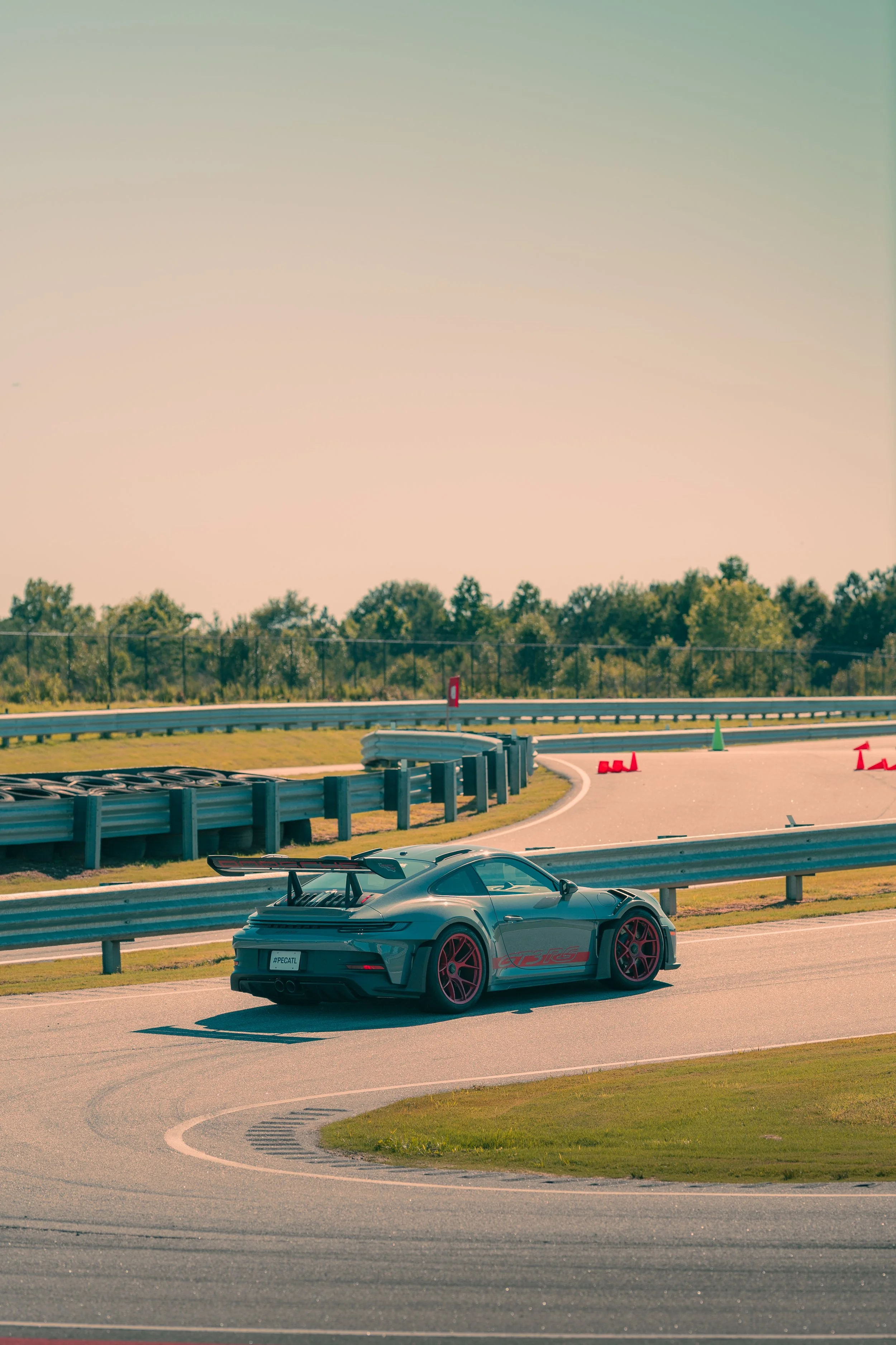 A sleek gray sports car with red accents and large rear wing parked on a race track, with safety cones and barriers, trees in the background, and a clear, slightly hazy sky.