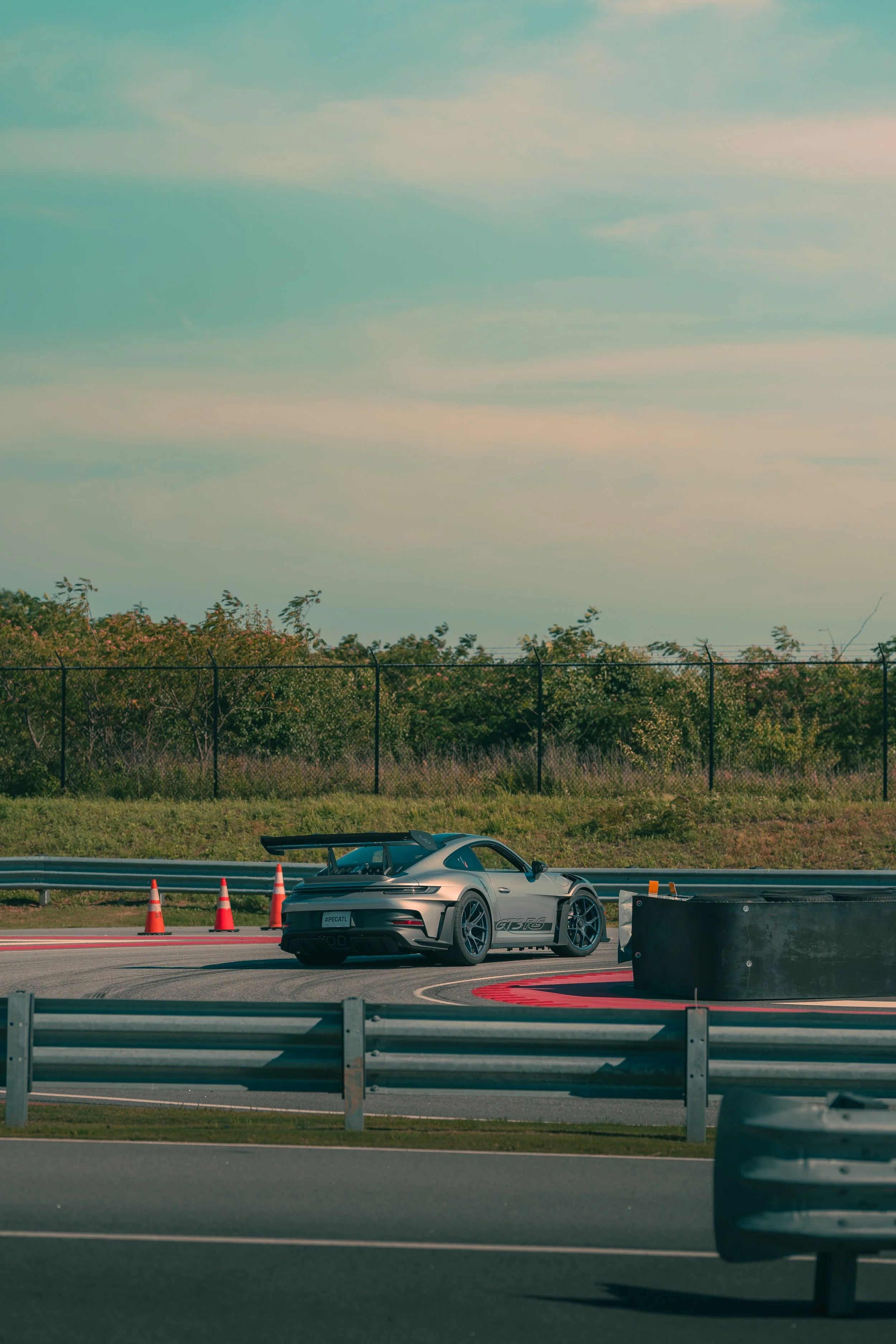 A silver Porsche 911 GT3 RS sports car on a racetrack with orange traffic cones and barriers, surrounded by safety barriers, with green bushes and trees in the background under a partly cloudy sky.