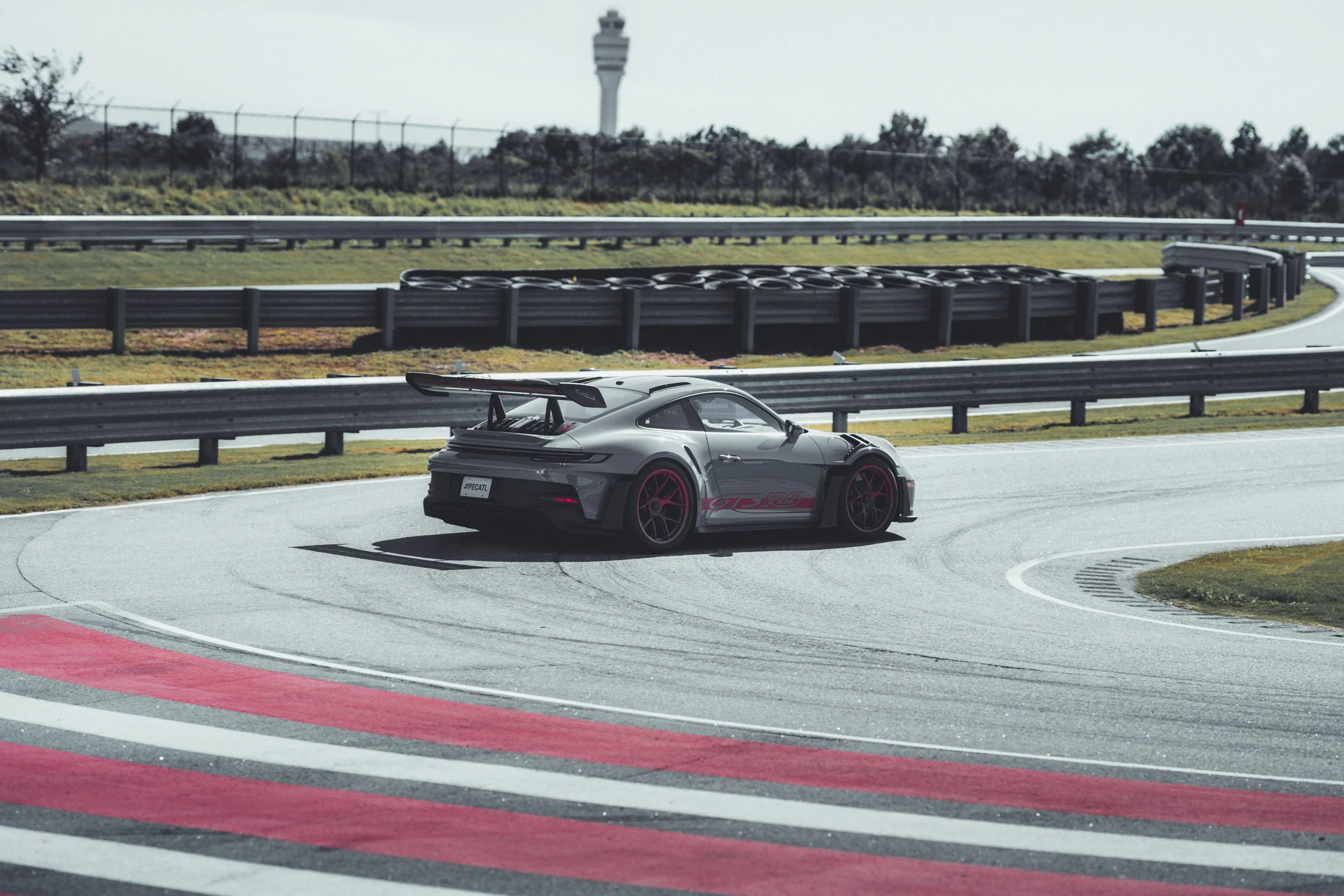 A gray sports car with a large rear spoiler on a racetrack, with a safety barrier and control tower in the background.