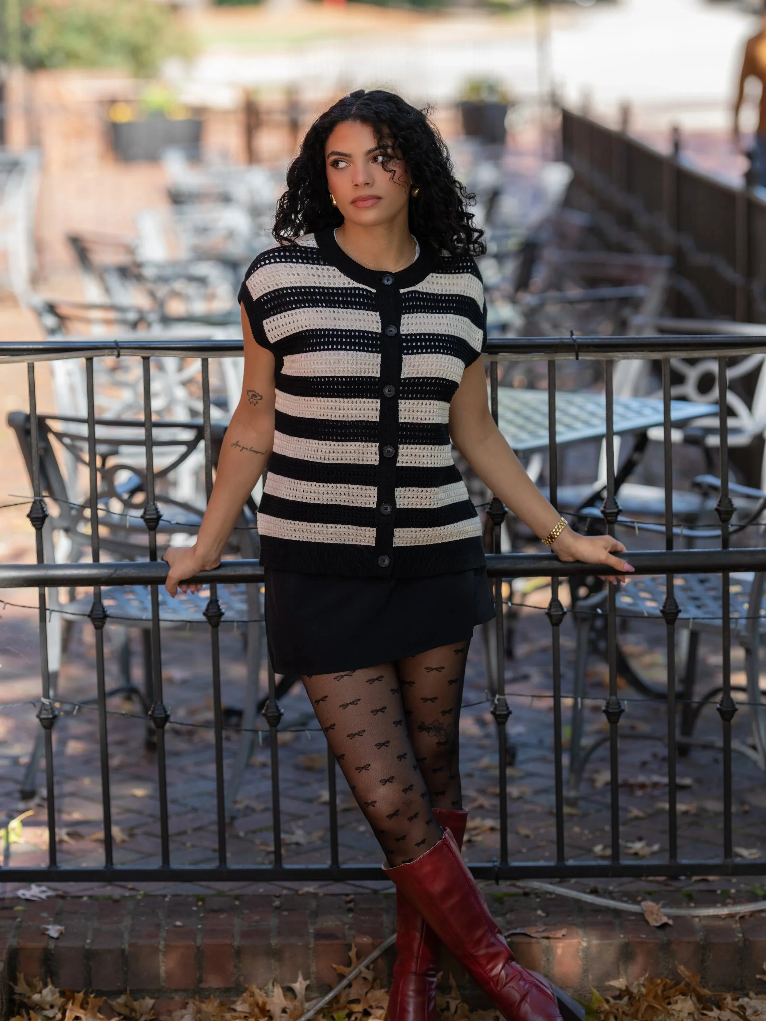 A young woman with dark, curly hair leaning on a black metal railing outdoors, wearing a black and beige striped buttoned vest, sheer tights with small bow patterns, red boots, and gold jewelry, with autumn leaves on the ground.