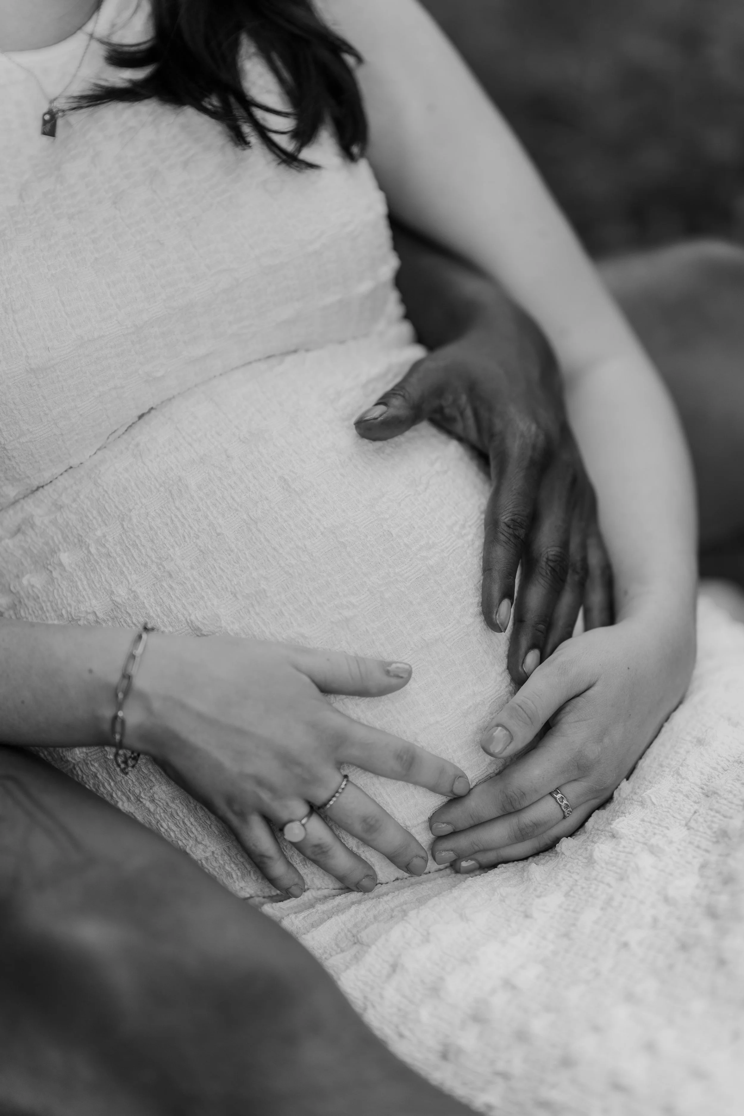 A person with dark hair and a light textured top has their hands on their pregnant belly, with multiple rings on their fingers and a bracelet on their wrist.