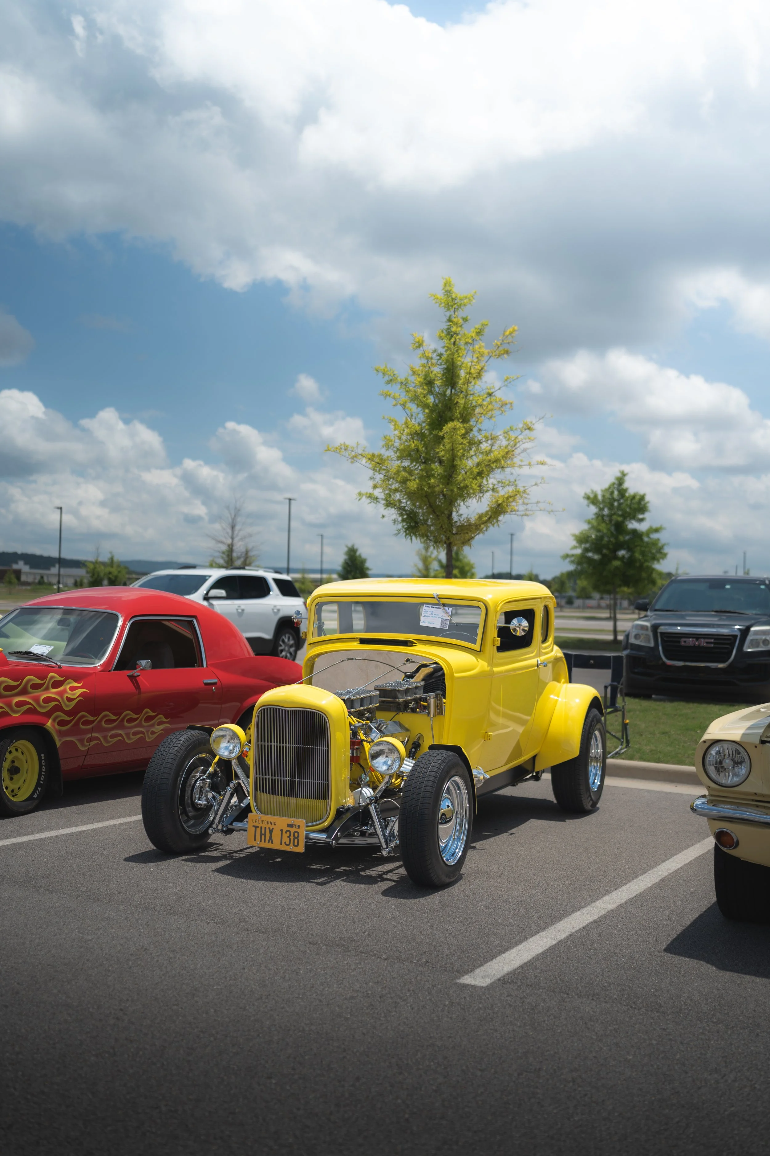 A yellow vintage hot rod car with an exposed engine on display at an outdoor car show, surrounded by other cars and trees under a partly cloudy sky.