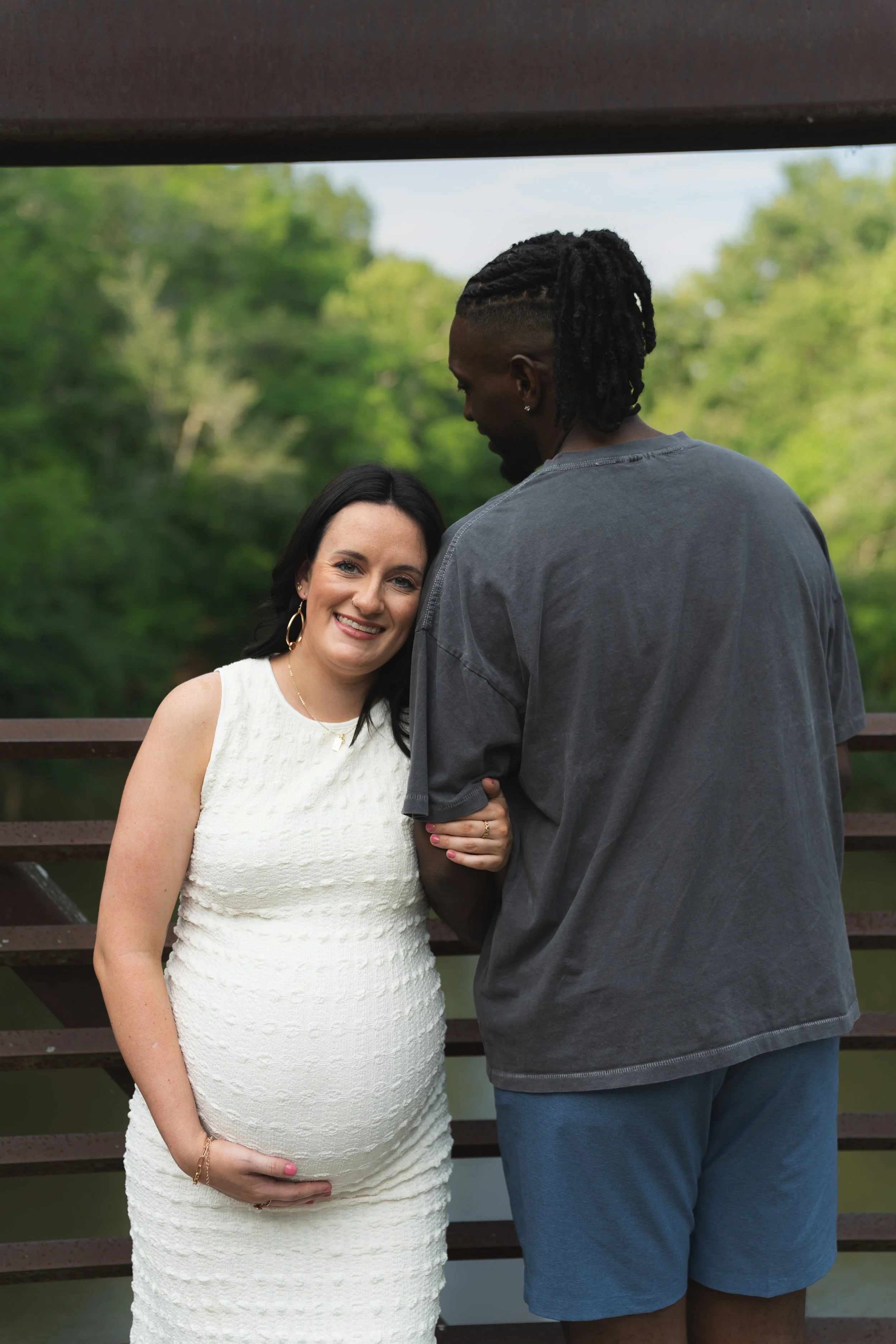 A pregnant woman with black hair smiling and wearing a white dress, and a man with battle rasta hairstyle, dark shirt, and blue shorts, standing outdoors on a wooden bridge with green trees in the background.