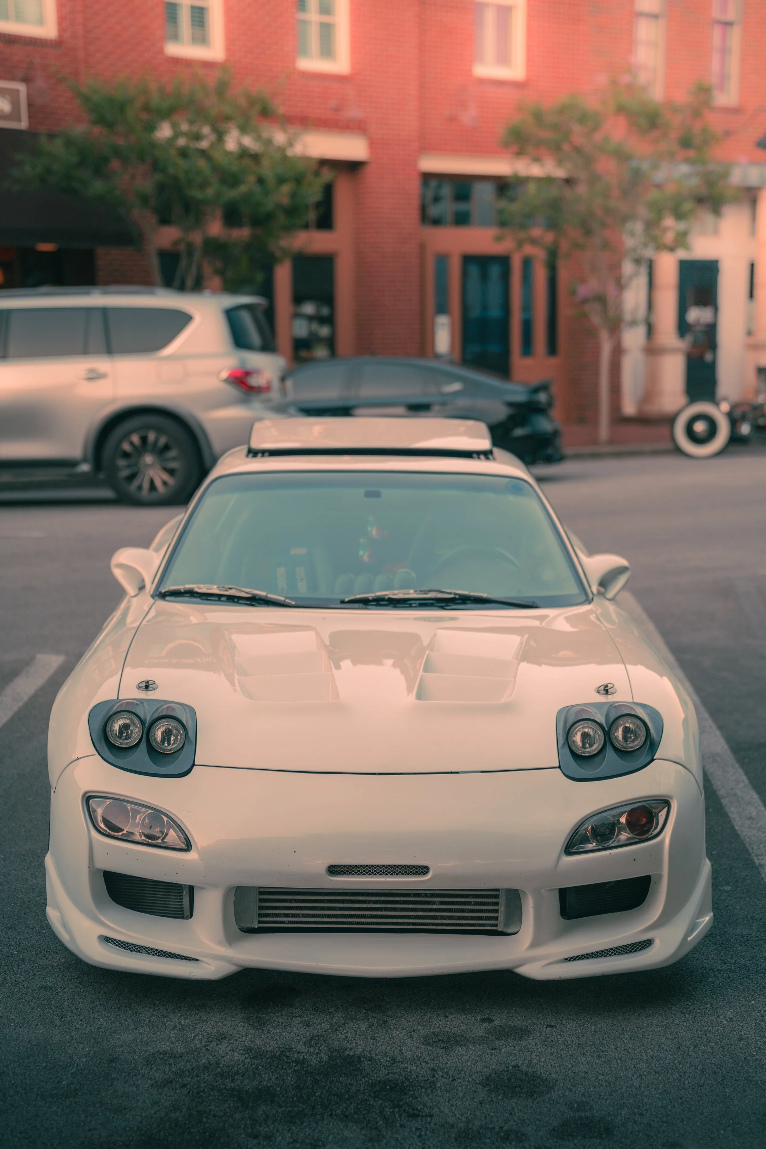 Front view of a white sports car parked on the street with other cars and a brick apartment building in the background.