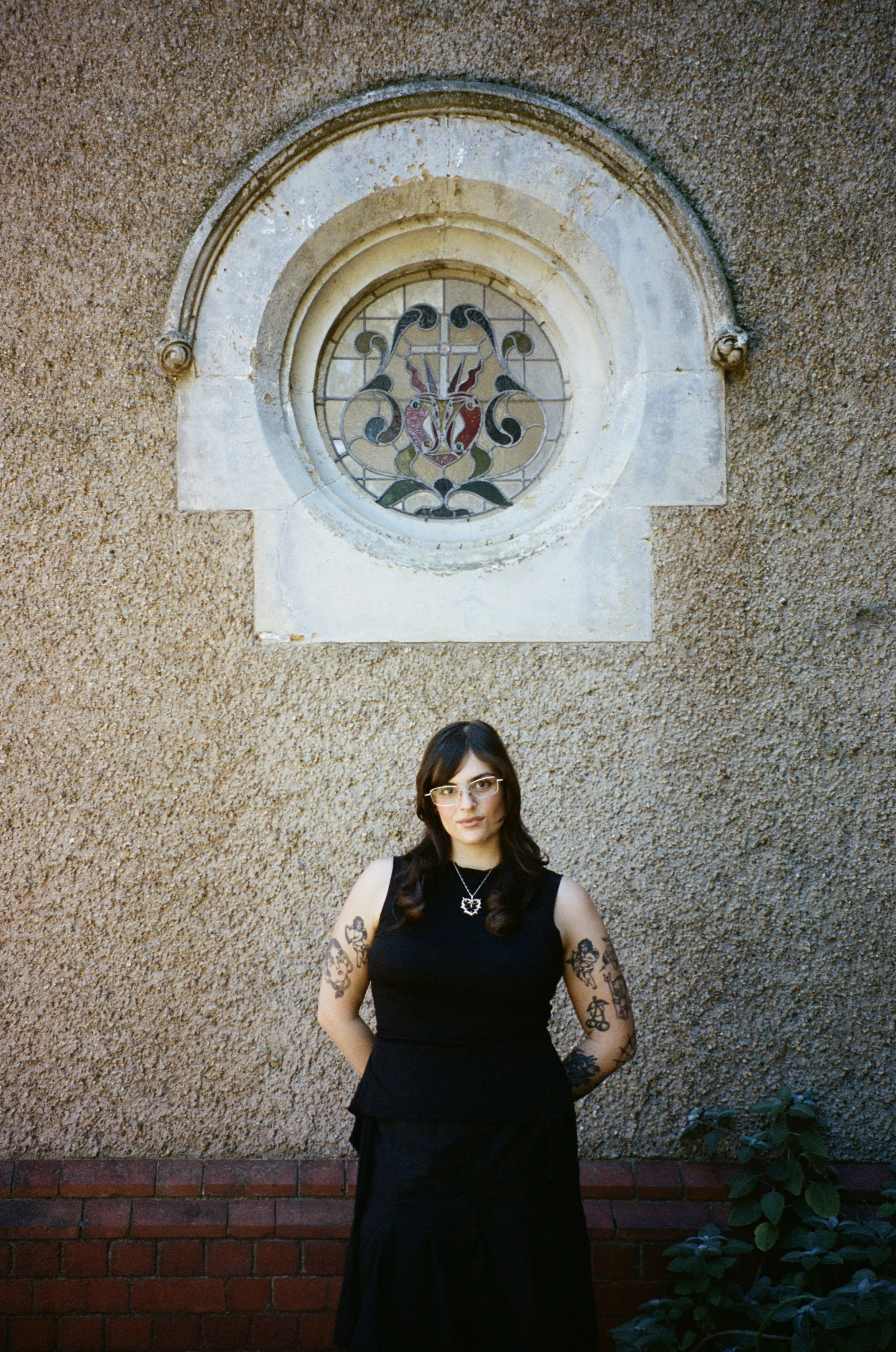 A woman with tattoos on her arms, wearing glasses and a black sleeveless top, standing outdoors in front of a textured beige wall with an arched stained glass window featuring a heraldic crest and decorative scrollwork.