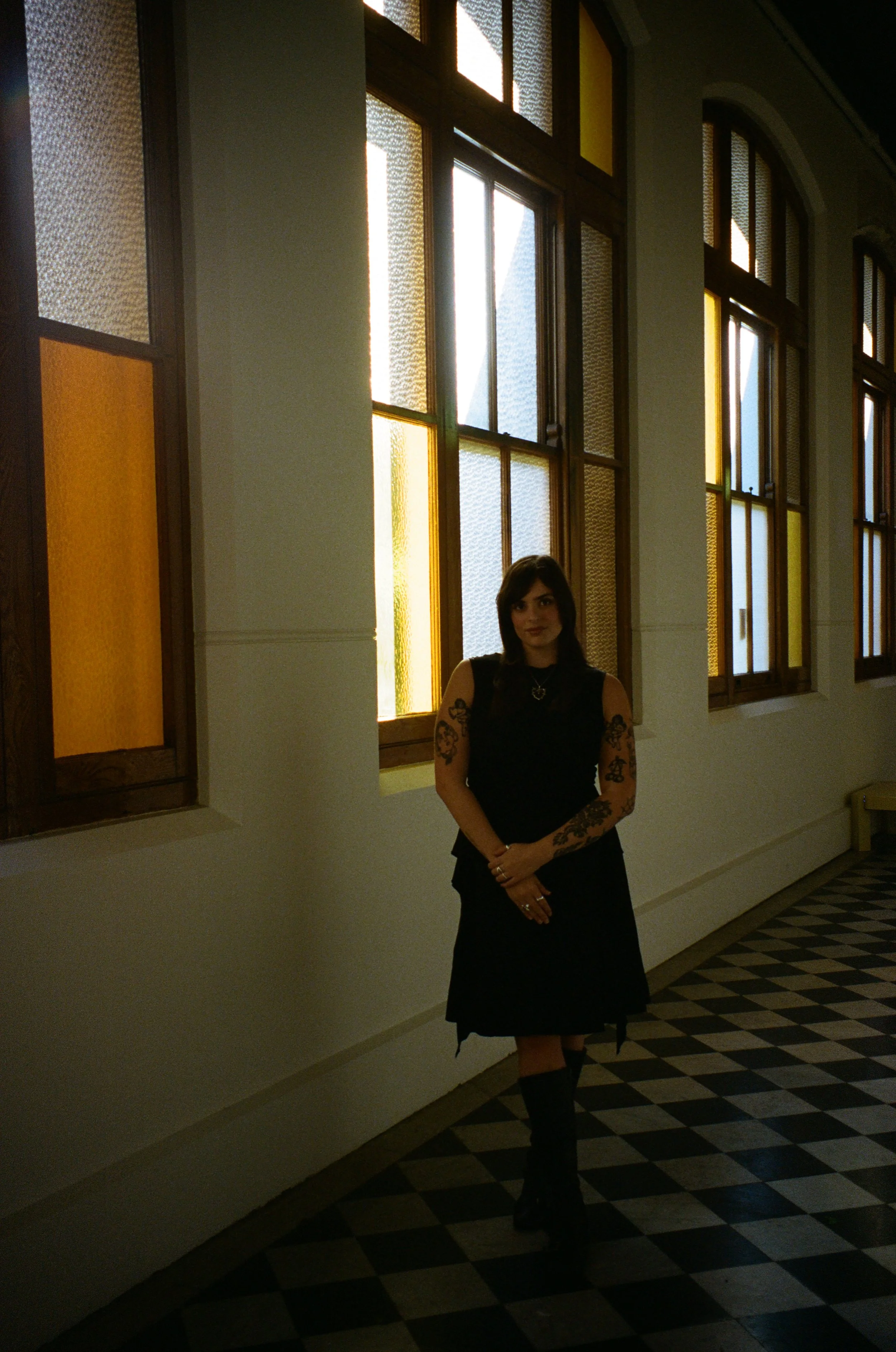 Woman with dark hair, tattoos on arms, wearing a black dress and knee-high boots, standing in a hallway with large stained glass windows and patterned floor.