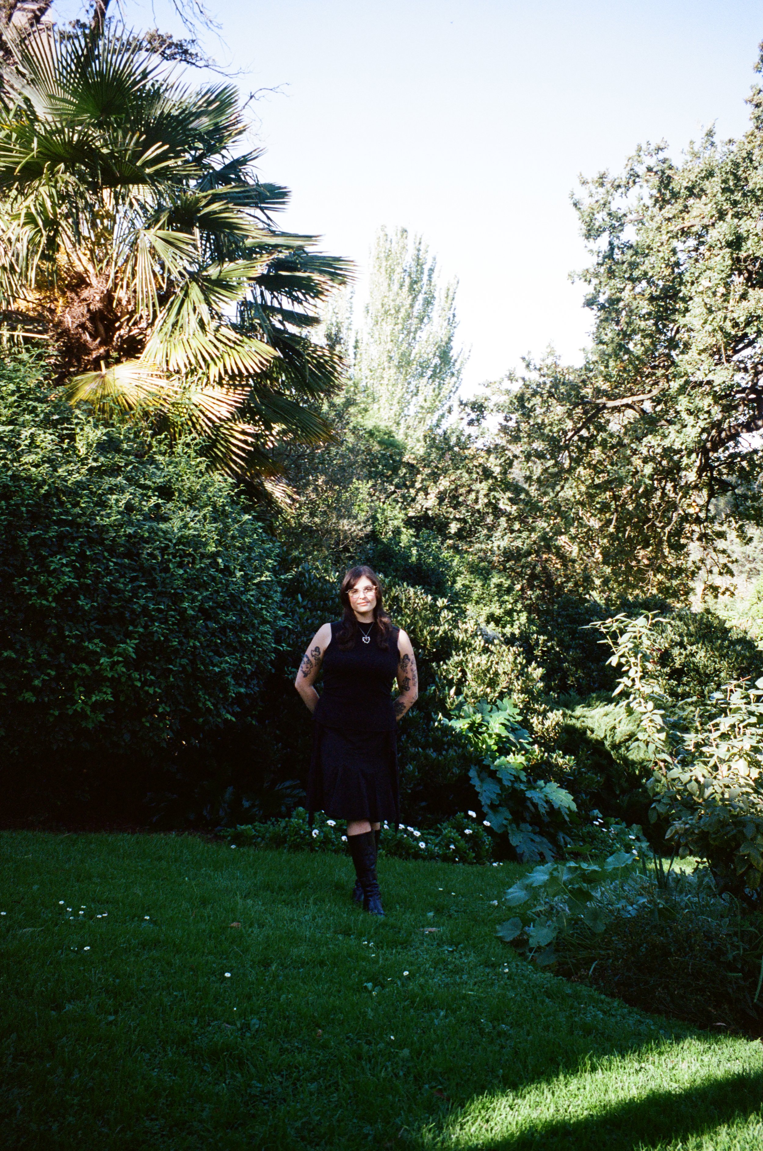 A woman with black hair and tattoos on her arms stands in a lush garden with various trees and bushes, wearing a sleeveless black top and a long black skirt. Bright sunlight illuminates the scene, creating shadows on the grass.