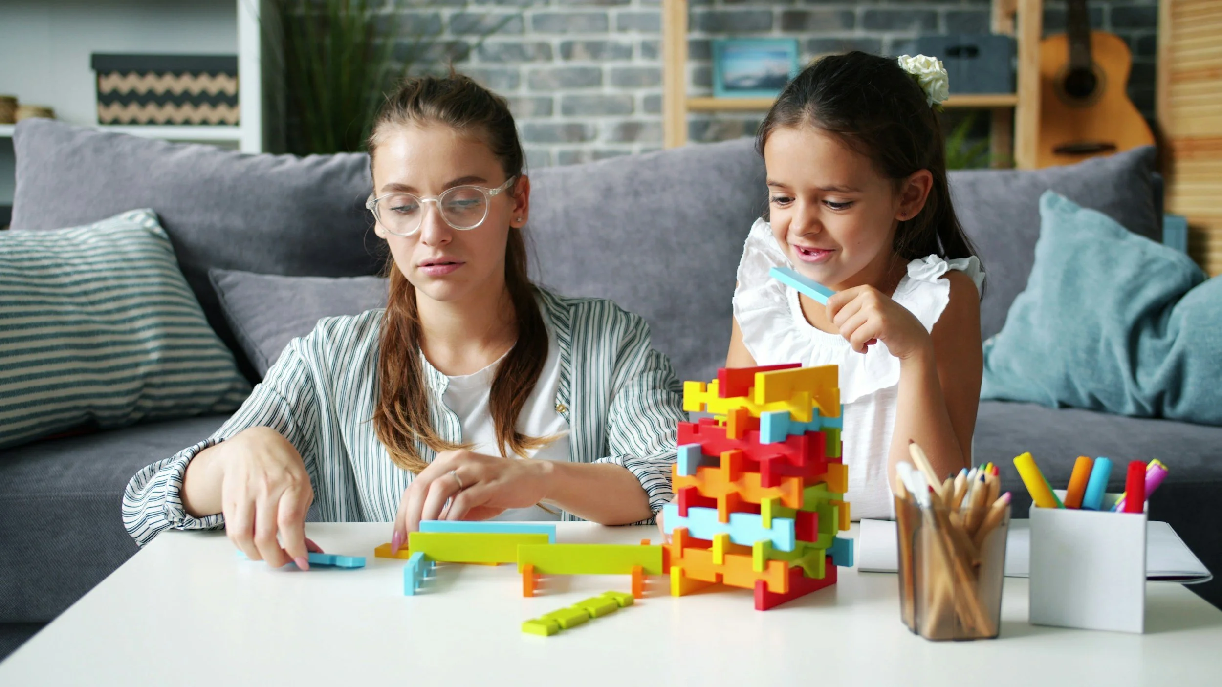 Two girls playing with colorful interlocking building blocks at a white table in a cozy living room.