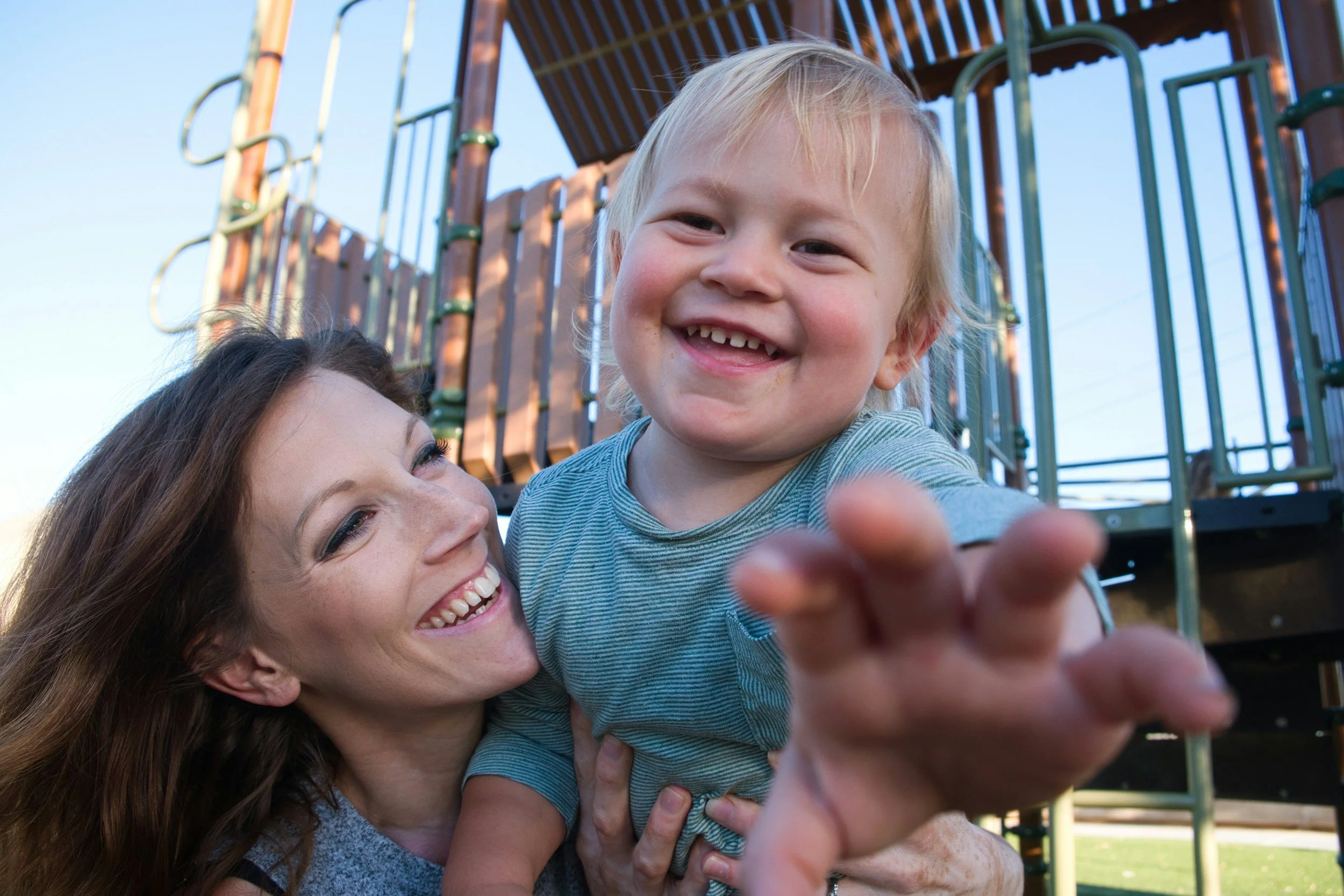 A smiling woman with long brown hair holding a happy young blonde boy on a playground