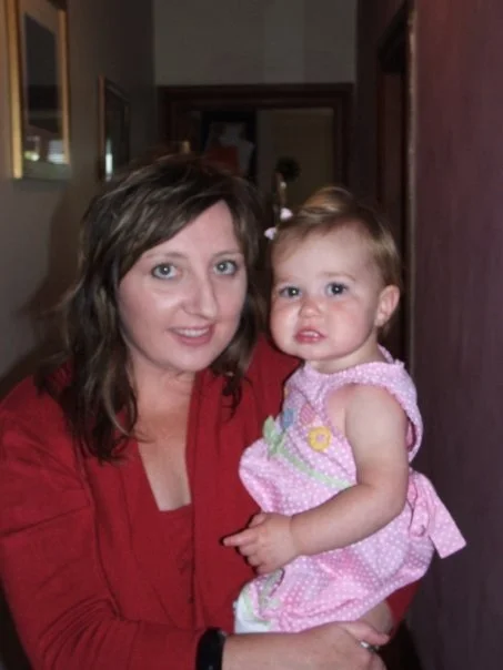 A woman holding a young girl, both looking at the camera in a home setting.
