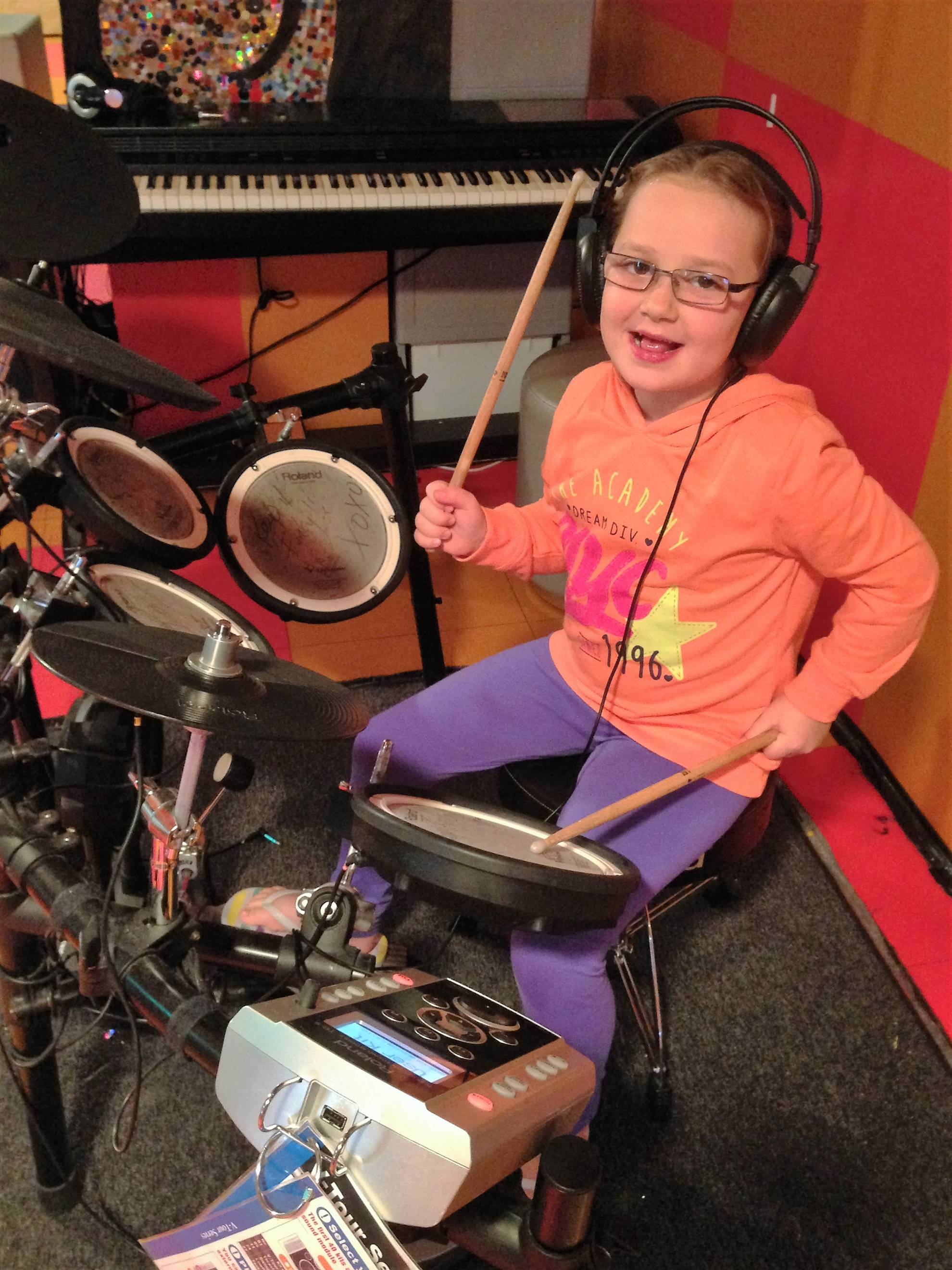 A young boy with glasses playing electronic drums, wearing headphones, in a colorful room with a piano and music equipment.