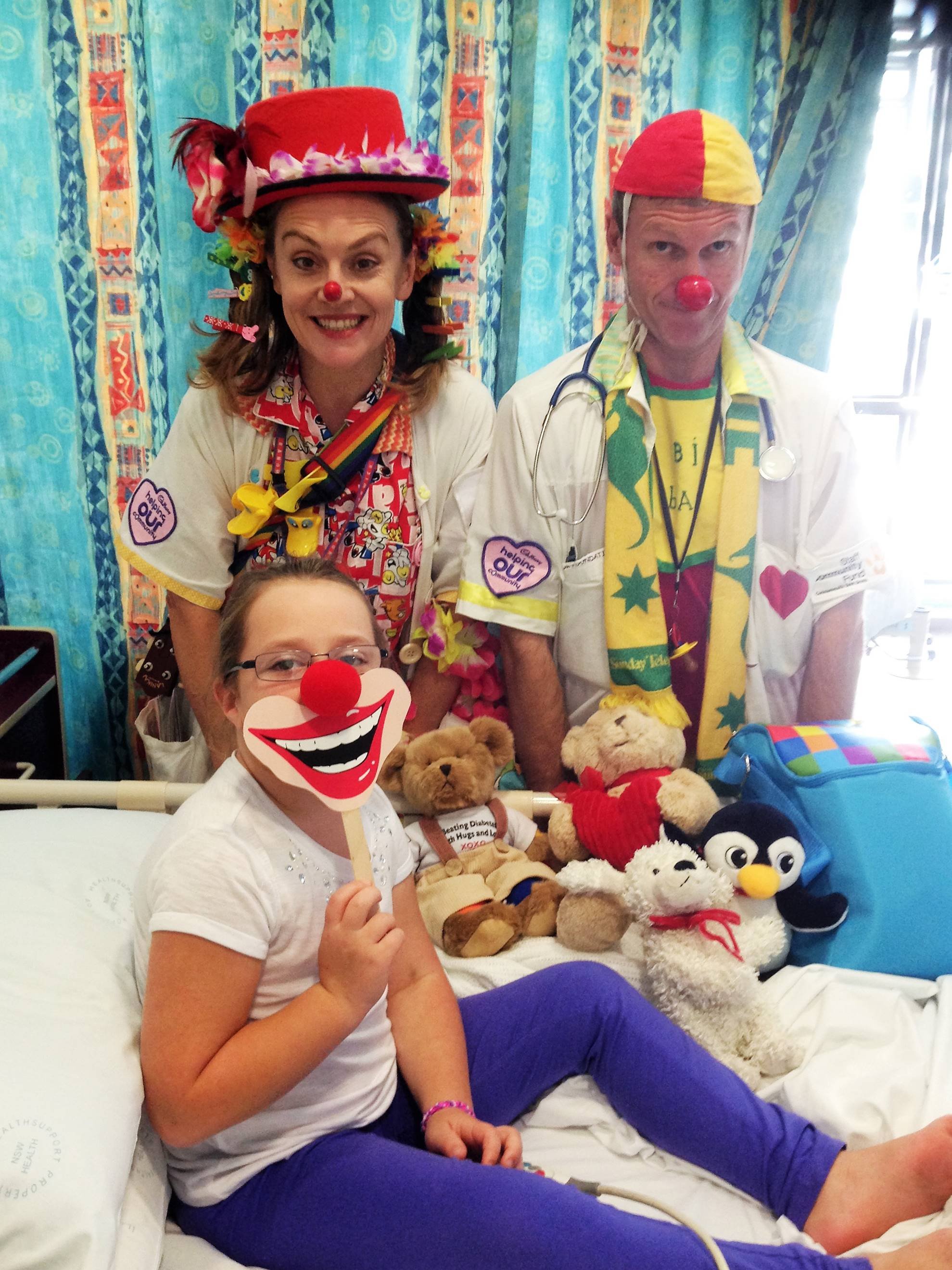 A group of people, including three adults and one child, dressed as clowns and medical staff, sitting on a hospital bed with teddy bears and plush toys, in a colorful room with patterned curtains.