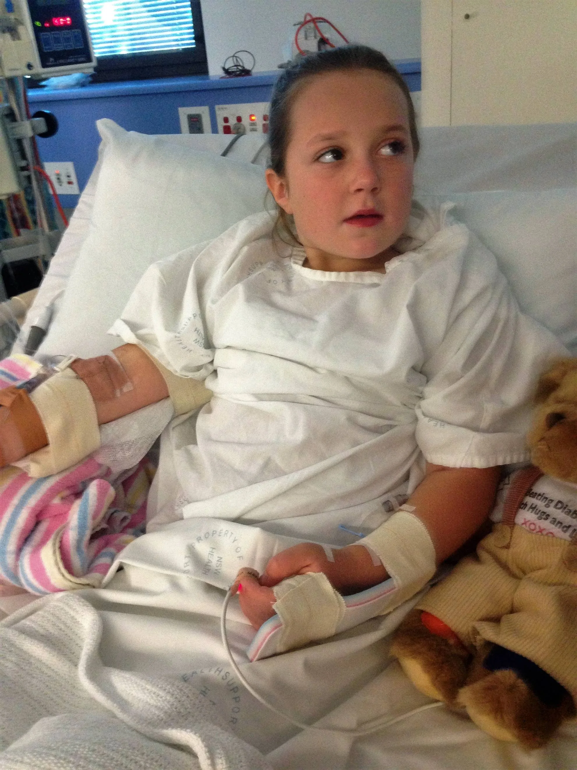 A young girl lying in a hospital bed with medical equipment and wires attached to her arm. She has dark hair tied back and is wearing a hospital gown. A small dog is lying beside her.