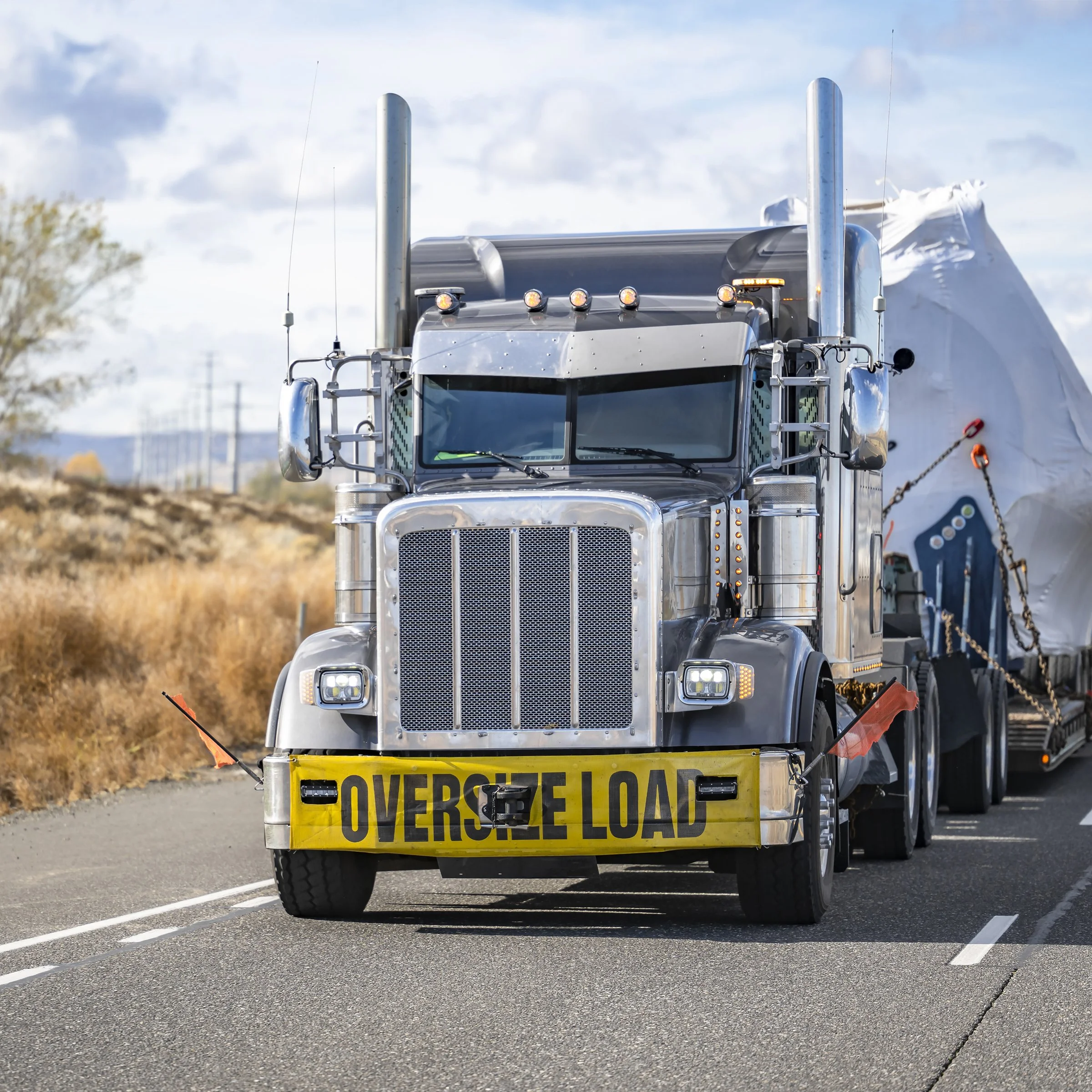 A semi-truck with a large yellow sign that reads 'OVERSEL LOAD' in black letters attached to the front bumper driving on a highway. The truck has a shiny silver grille, multiple light bars on top, and is hauling a large white load, possibly a generator or piece of equipment. The background shows a partly cloudy sky and some trees and utility poles along the road.