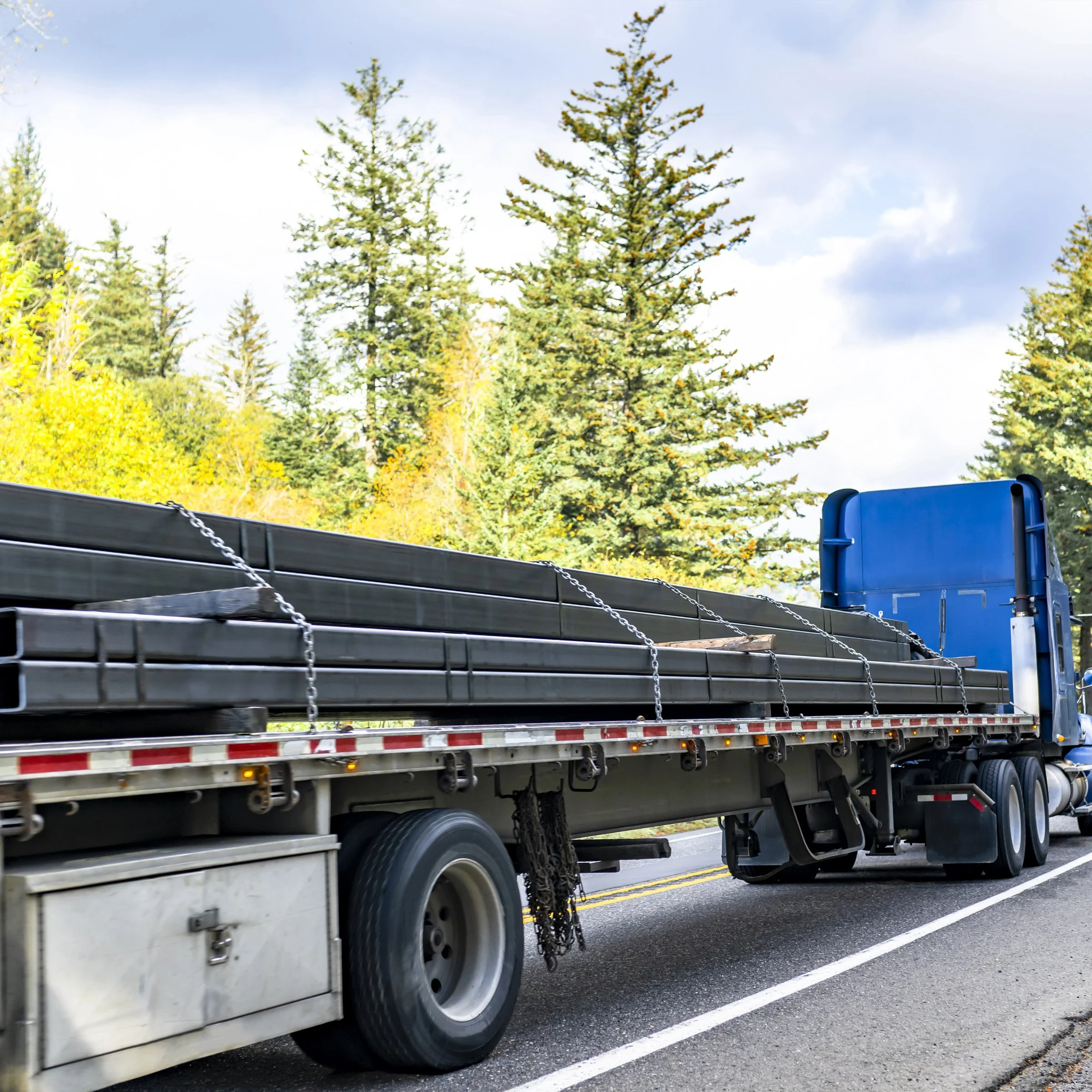 A flatbed truck carrying long metal pipes down a paved road with trees and yellow foliage in the background.
