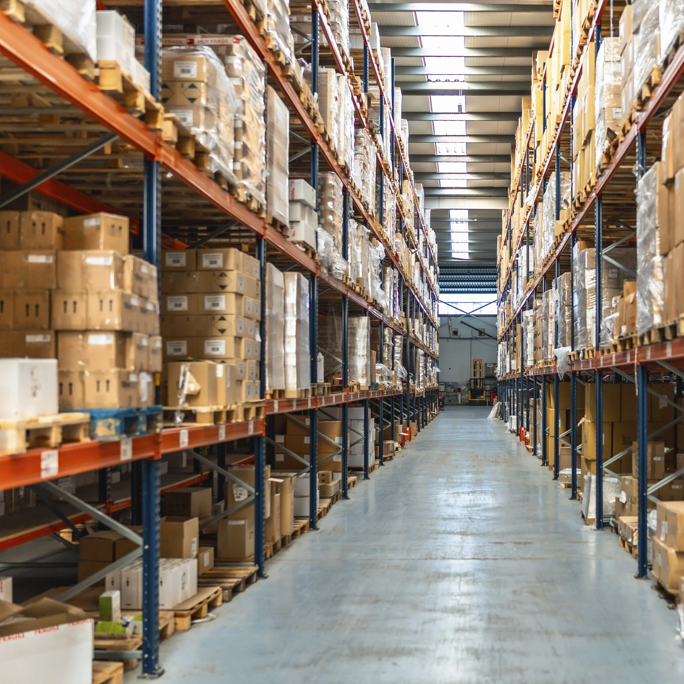 A warehouse aisle with tall shelves stocked with boxes and pallets, illuminated by skylights on the ceiling.