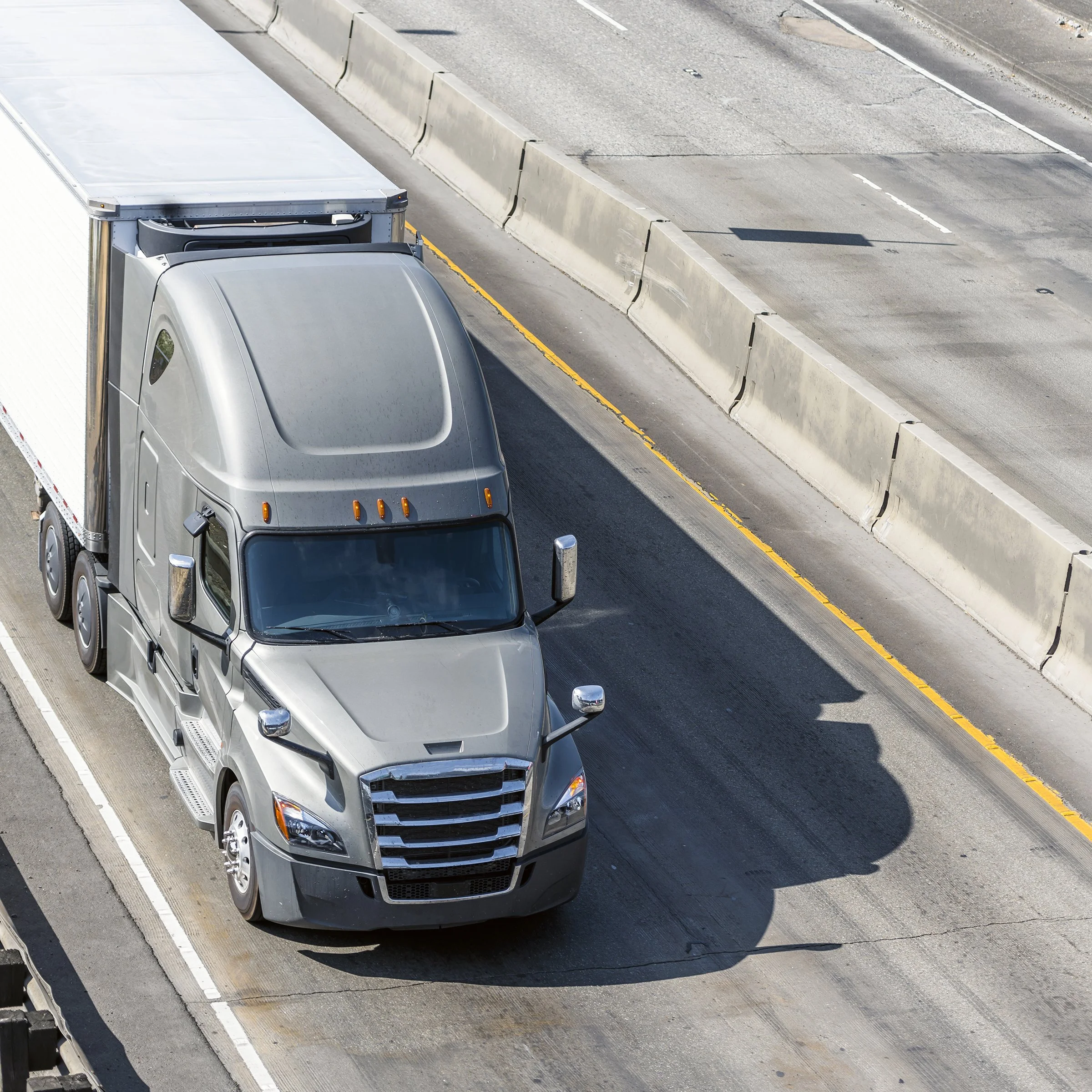 A gray semi-truck on a highway lane near concrete barriers with lane markings visible.