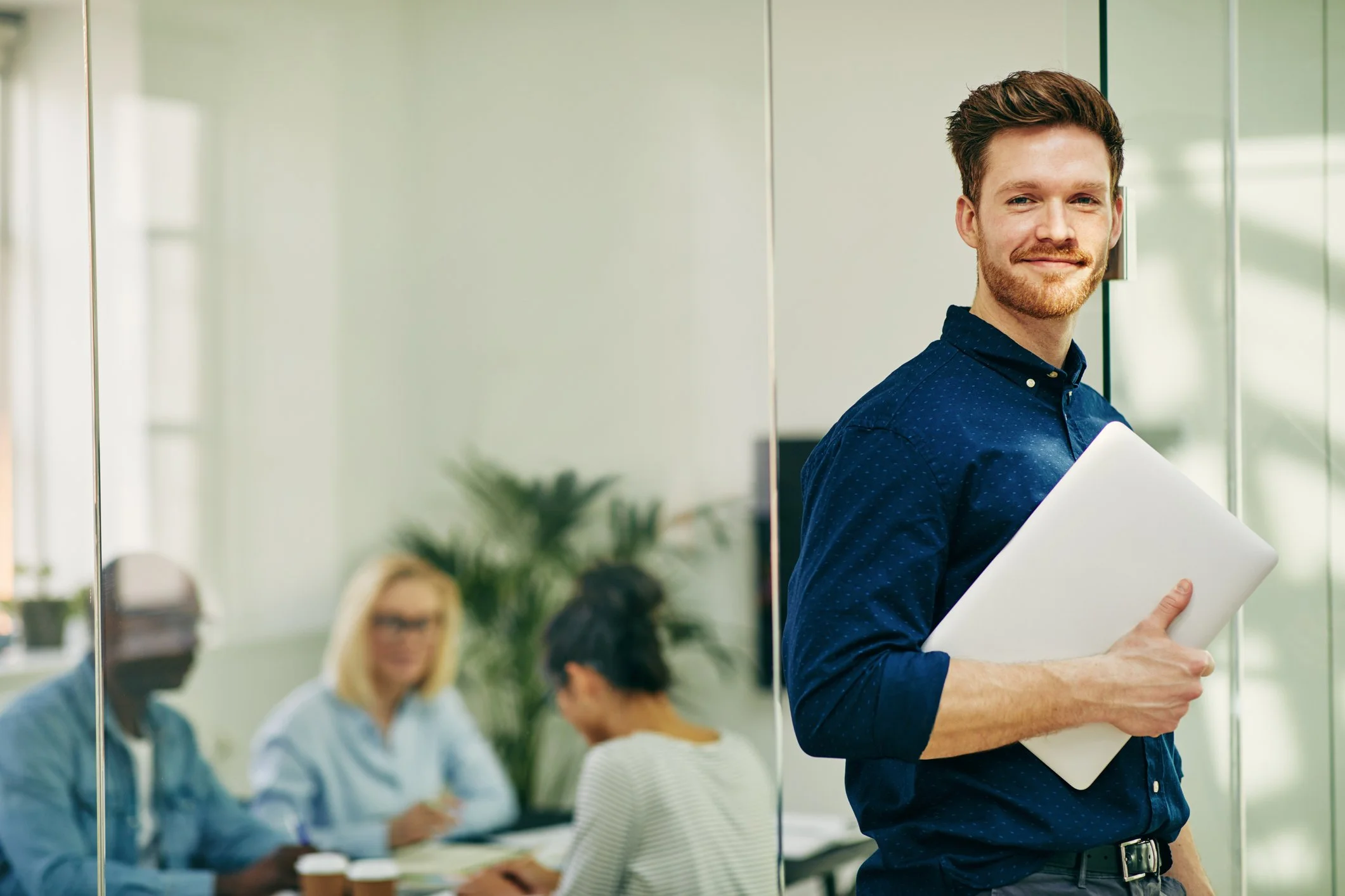 A young man with brown hair and a beard, wearing a blue shirt, holds a white paper or folder and smiles while standing in an office. In the background, two women are seated at a table, blurred, with one wearing glasses and a blue shirt, and the other blonde with striped shirt, in a modern office setting with green plants and glass walls.