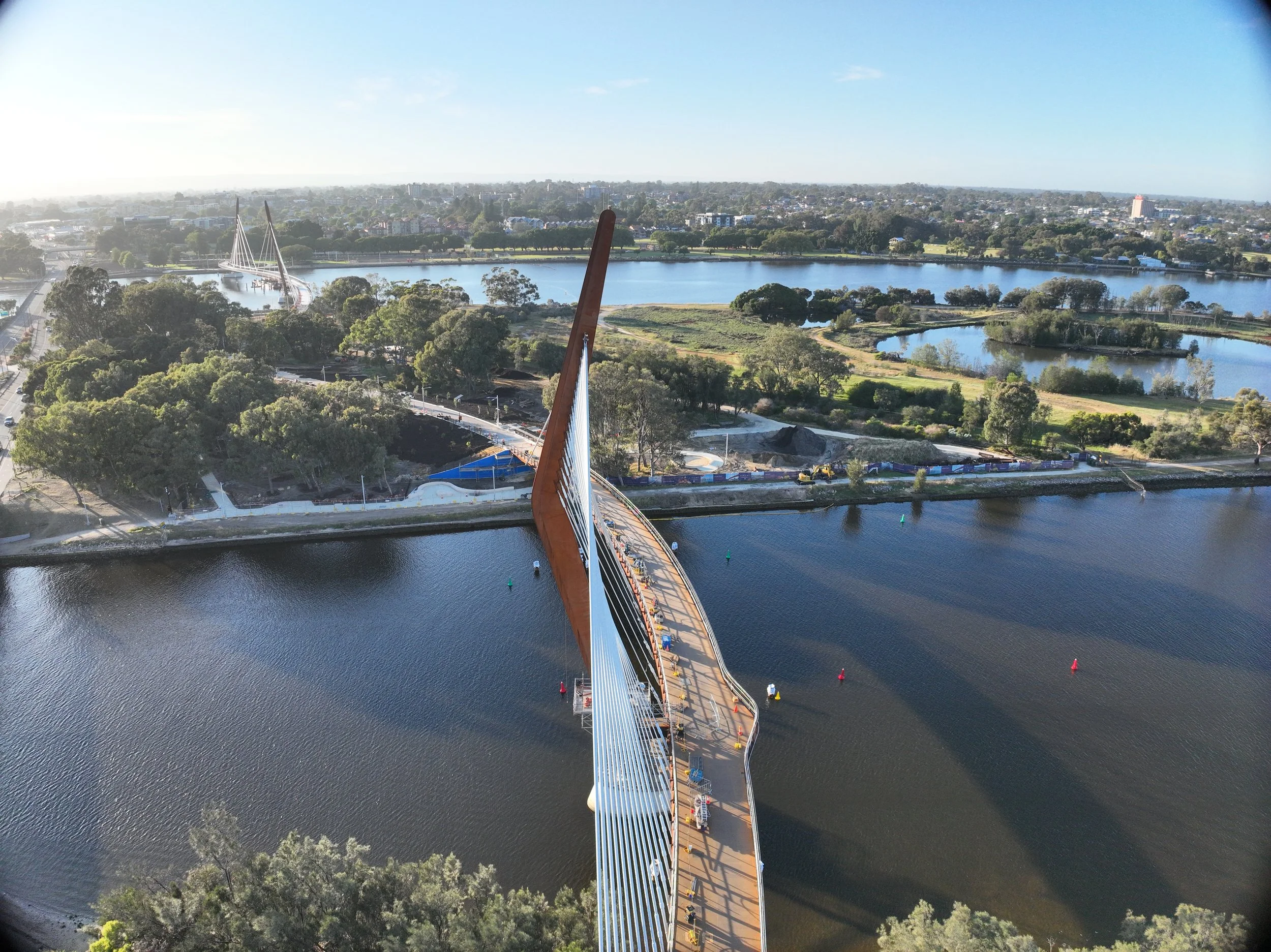 An aerial view of a long bridge crossing over a river, with a city skyline and green parks in the background under a partly cloudy sky.