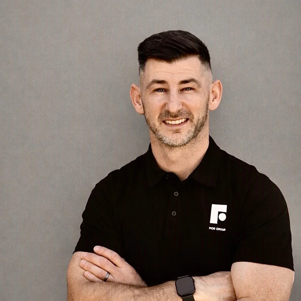 A smiling man with short dark hair and a beard, wearing a black polo shirt with a logo on the chest, standing against a gray background.