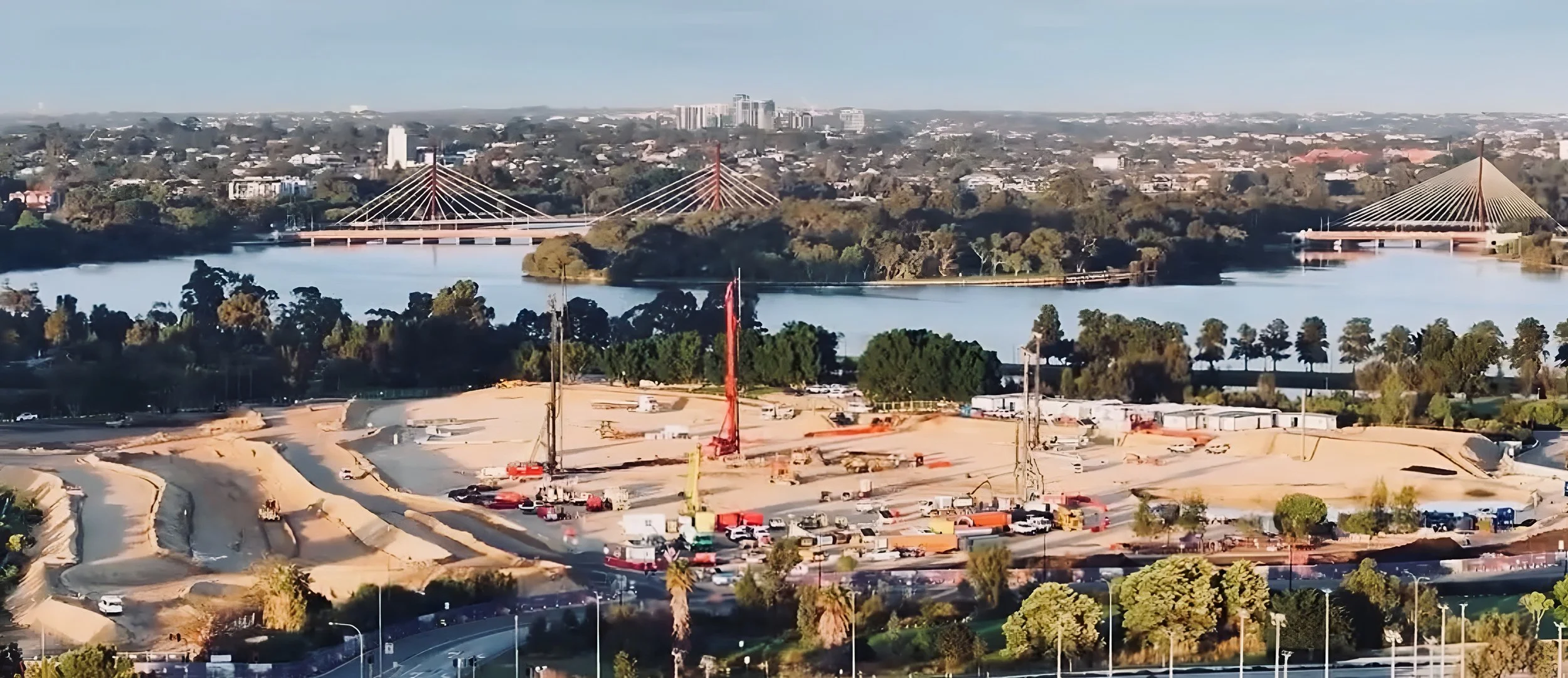 Construction site with dirt roads, vehicles, and cranes in the foreground, a lake with two bridge structures and bridges in the middle, and a cityscape with numerous buildings and trees in the background.