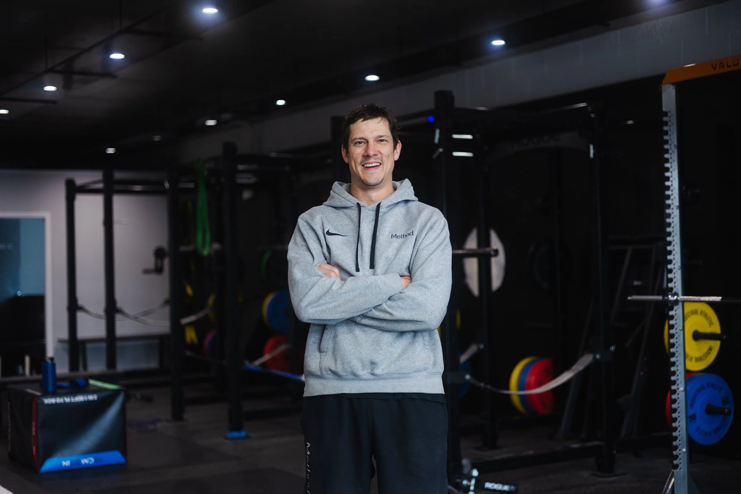 Man standing in a gym with arms crossed, smiling, wearing a gray hoodie with 'Method' logo, surrounded by workout equipment, black ceiling with spotlights.