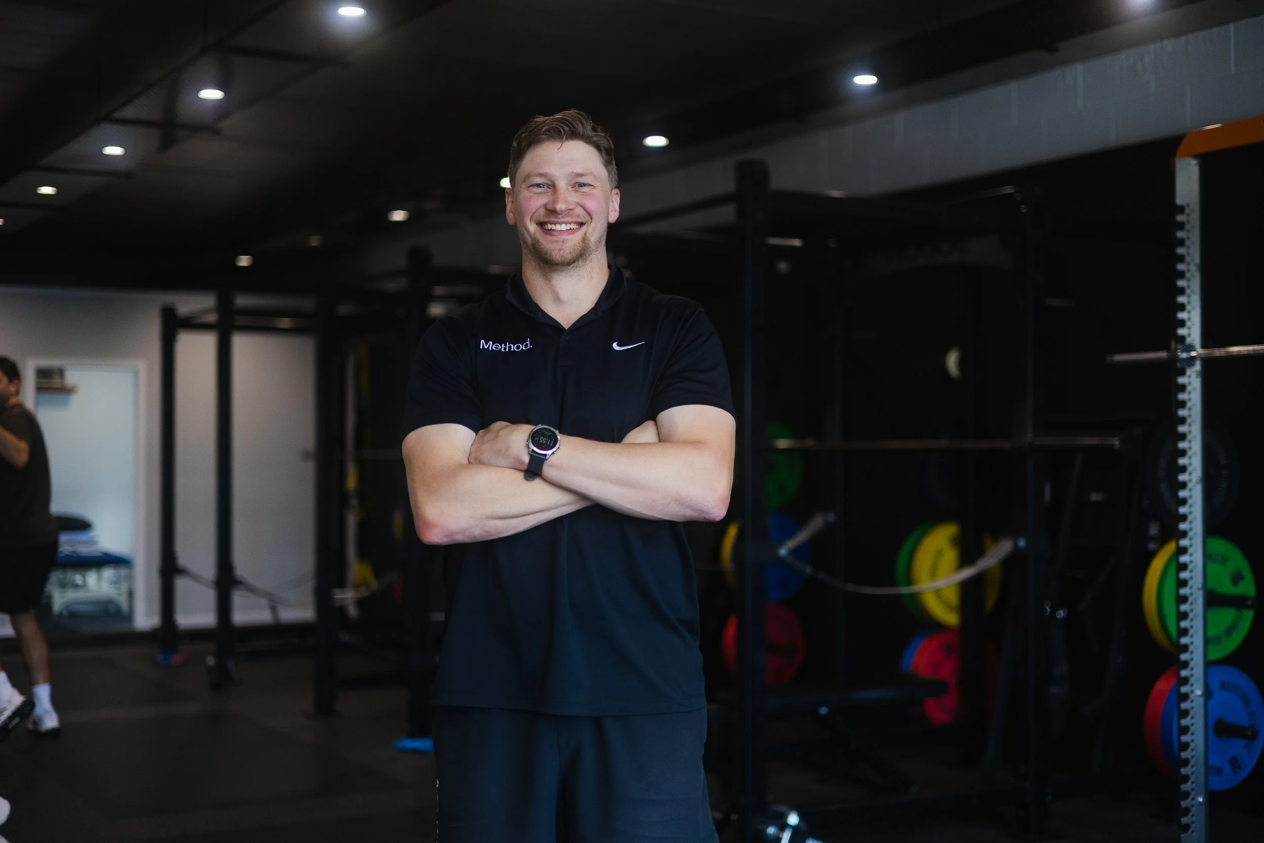 A young man with short hair and a beard smiling confidently at the gym, wearing a black sports shirt with "Method", standing with arms crossed in front of weightlifting equipment.