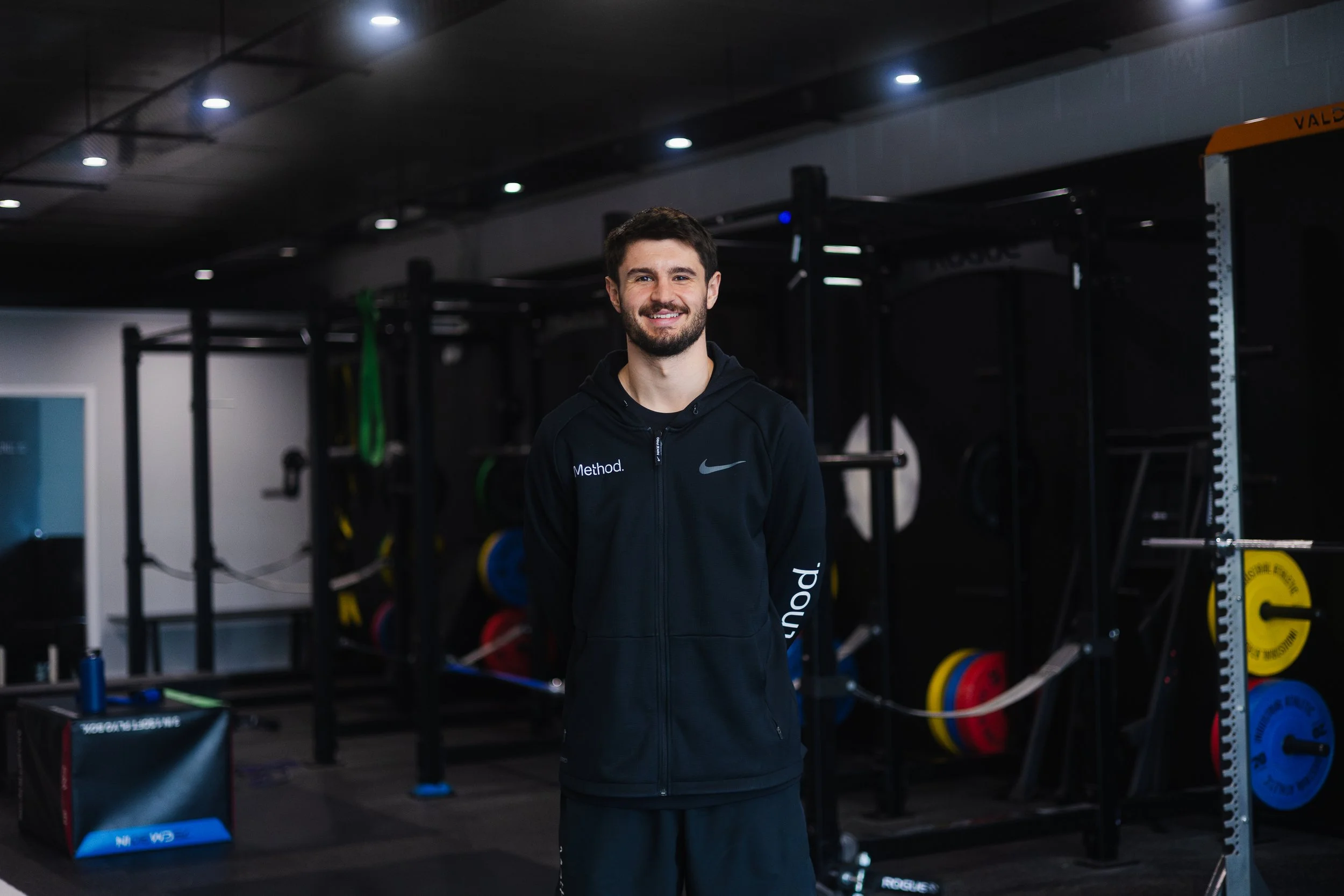 Smiling man in black athletic wear standing in a gym with weightlifting equipment, including barbells, weights, and gym accessories, in the background.