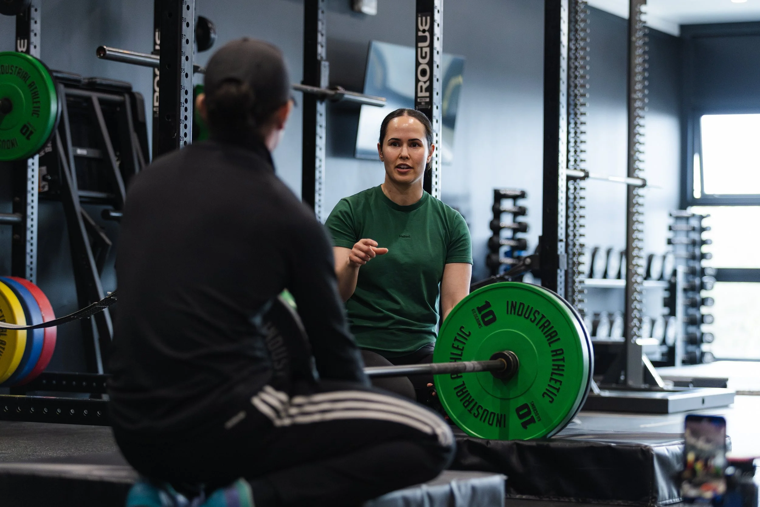 A woman in a green shirt talking to a man in a gym, with weightlifting equipment and weights in the background.