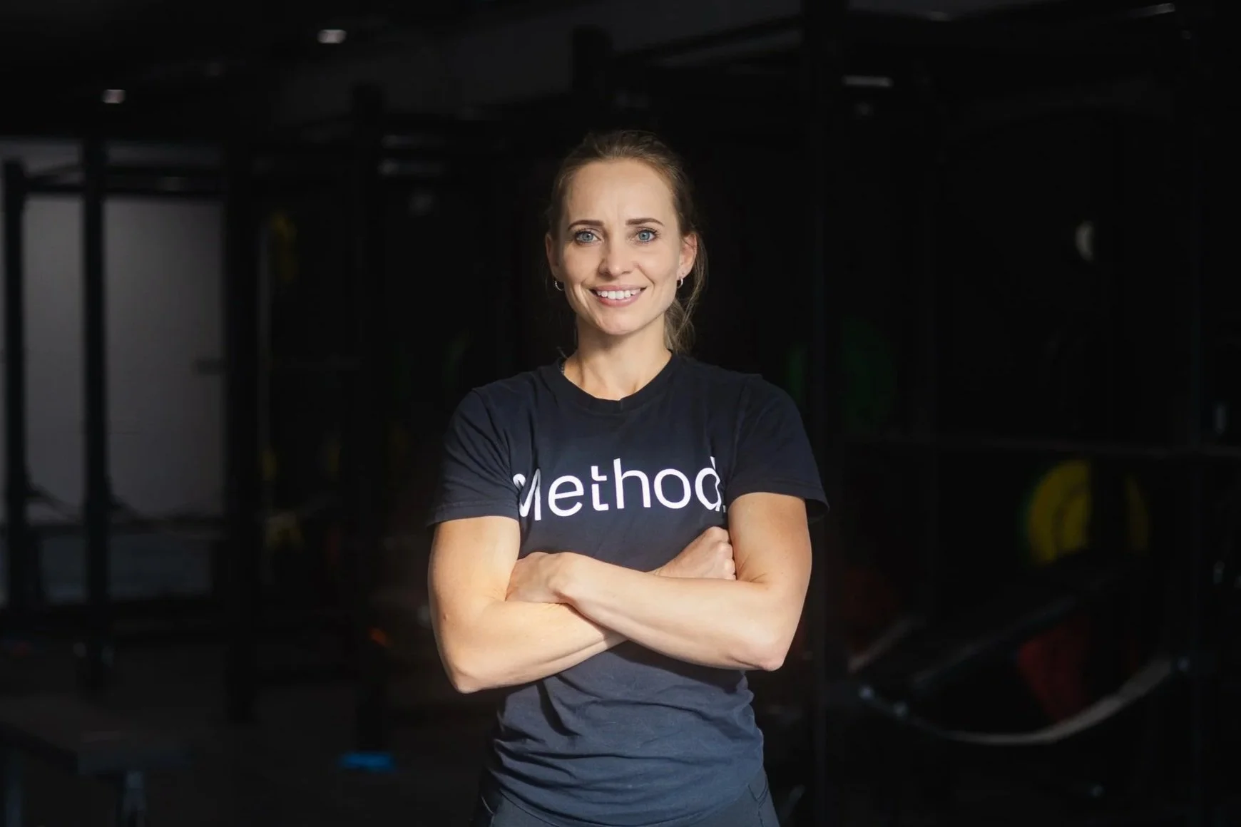 A confident woman with crossed arms smiling at the camera in a gym setting, wearing a black t-shirt with the word 'Method' on it.