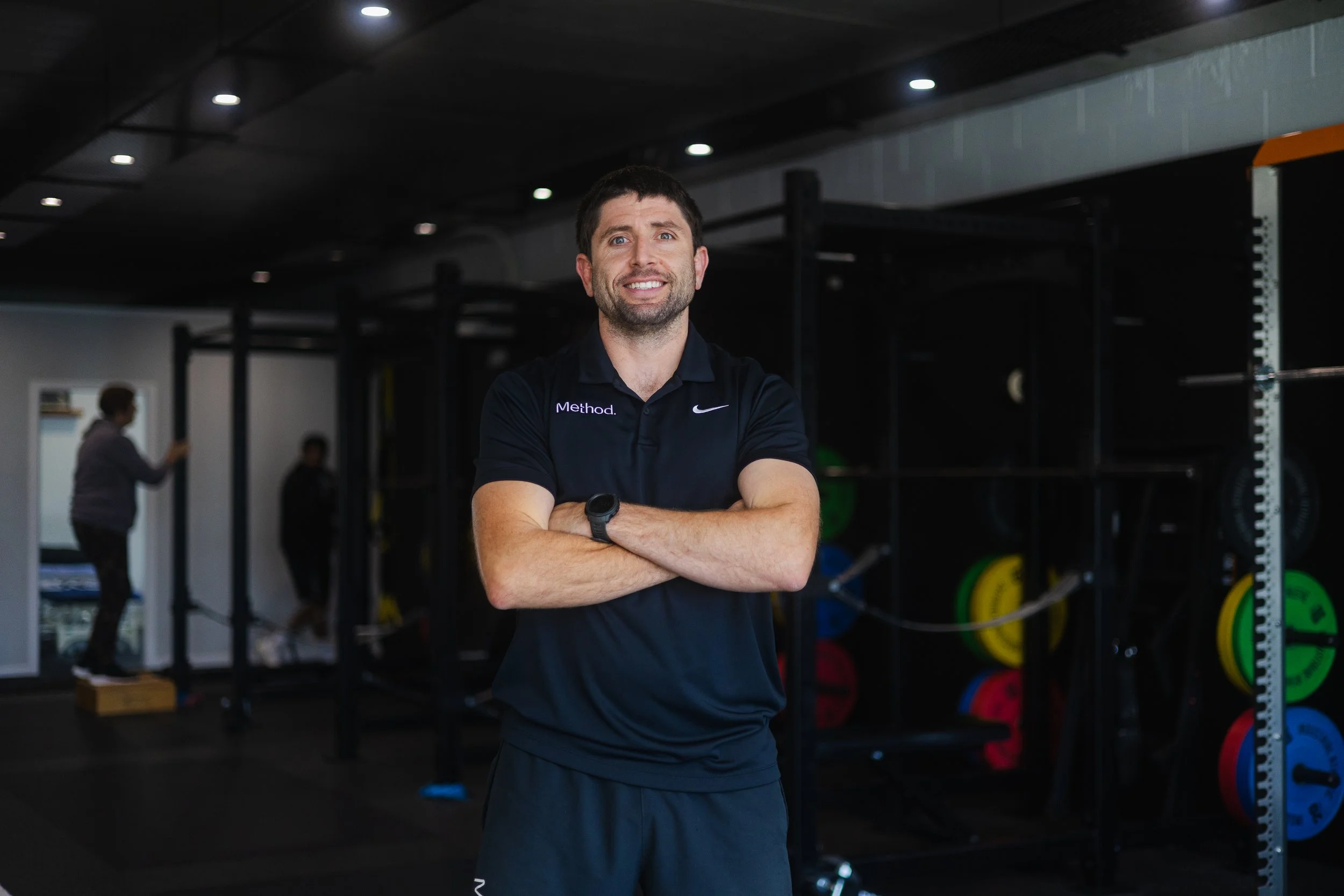 A man with dark hair and a beard, wearing a black sports shirt with the word "Method", stands with arms crossed in a gym with weightlifting equipment and colorful weight plates in the background.