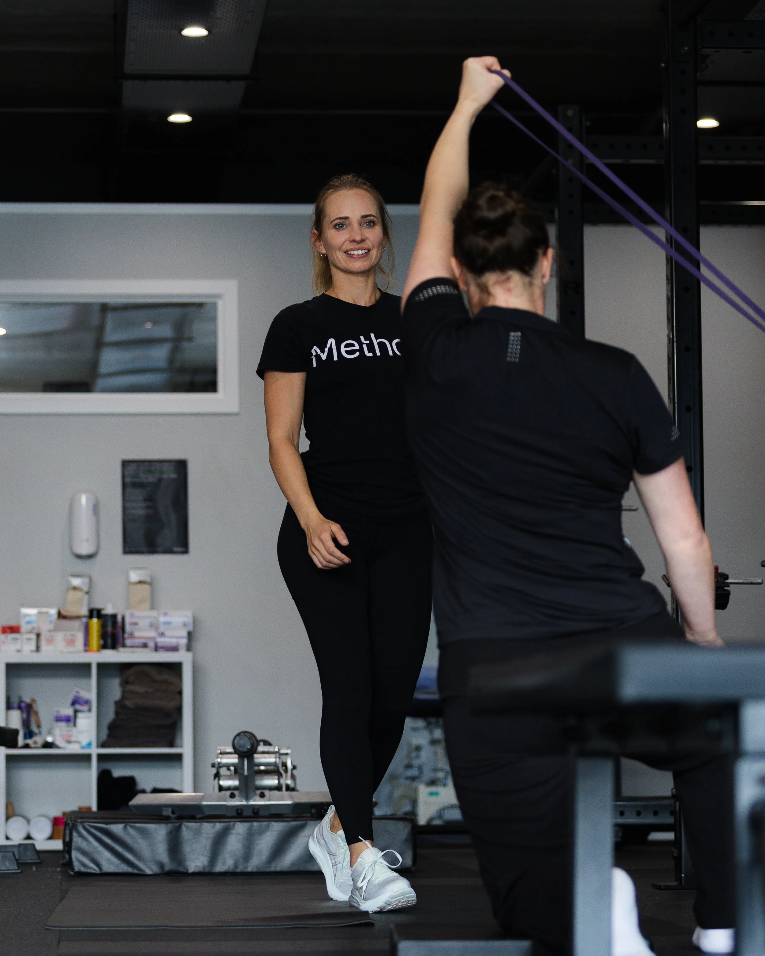 Two women exercising in a gym, one stretching her leg while the other observes, with workout equipment and shelves in the background.