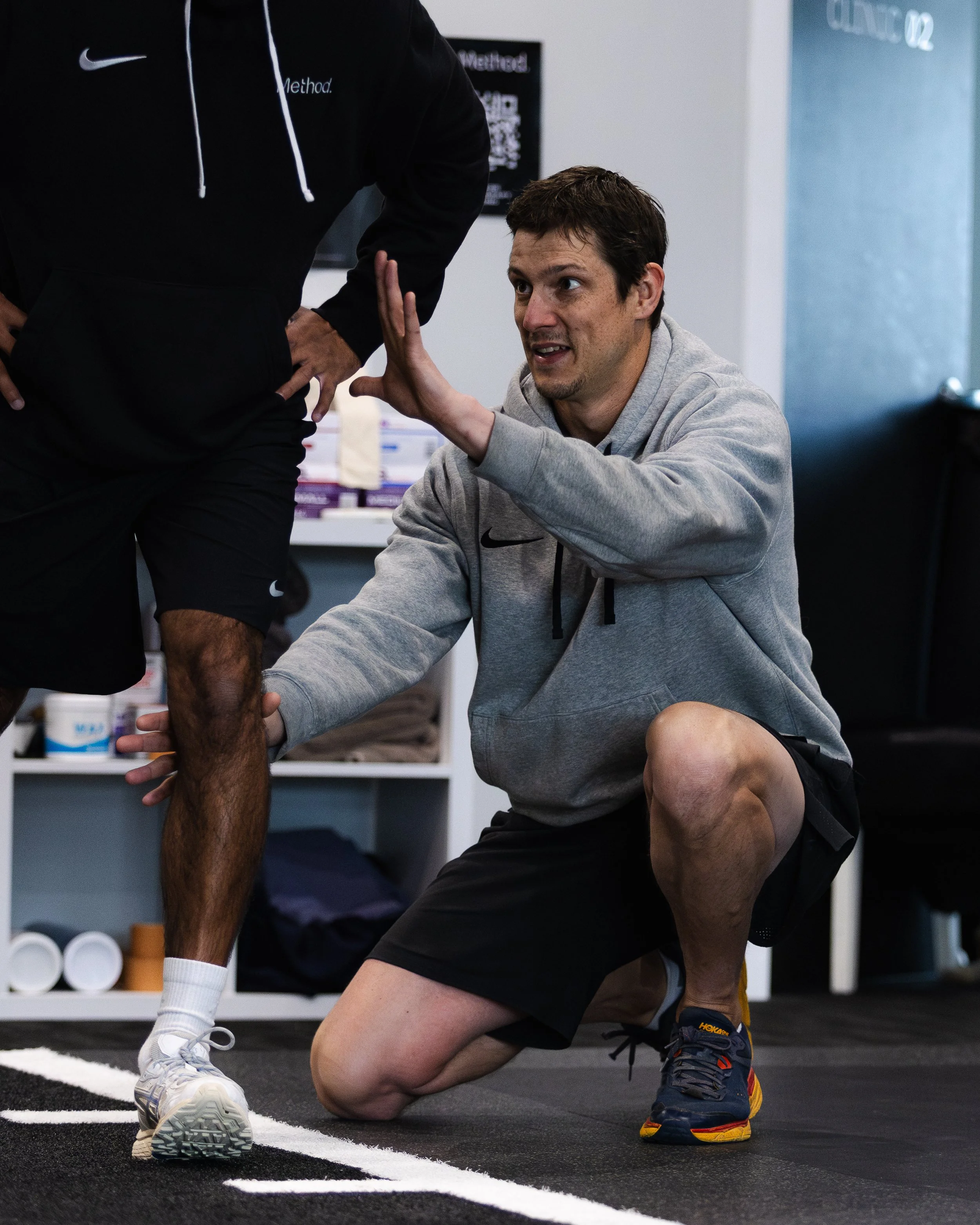 A man in a gray hoodie kneeling on one knee on a gym floor, checking a runner's leg. The runner is wearing black shorts, a black hoodie, and white shoes, with a person standing nearby.