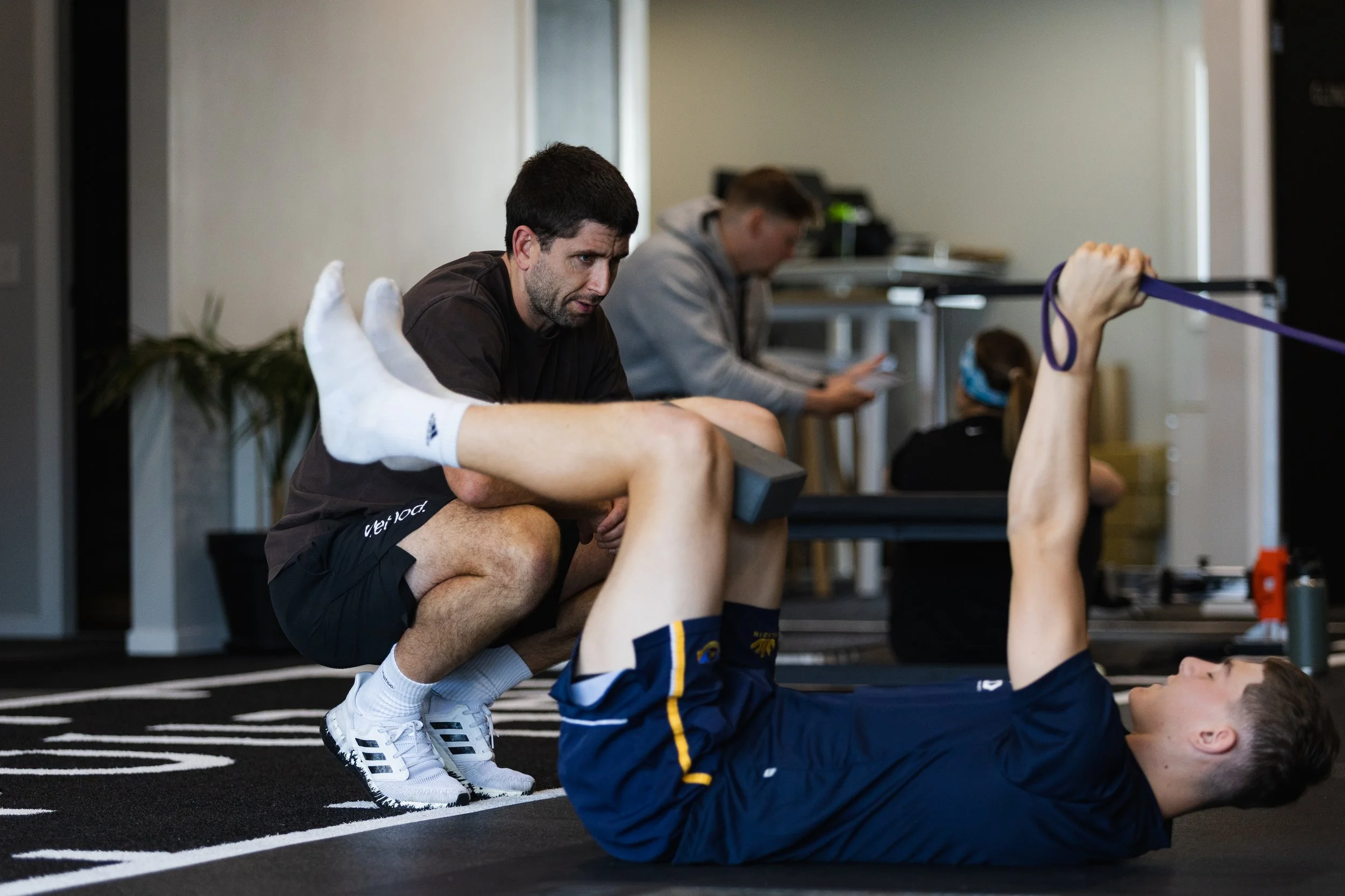 A man assisting a client with an exercise at a gym, with the client lying on a mat and lifting their legs while the man supports their back.