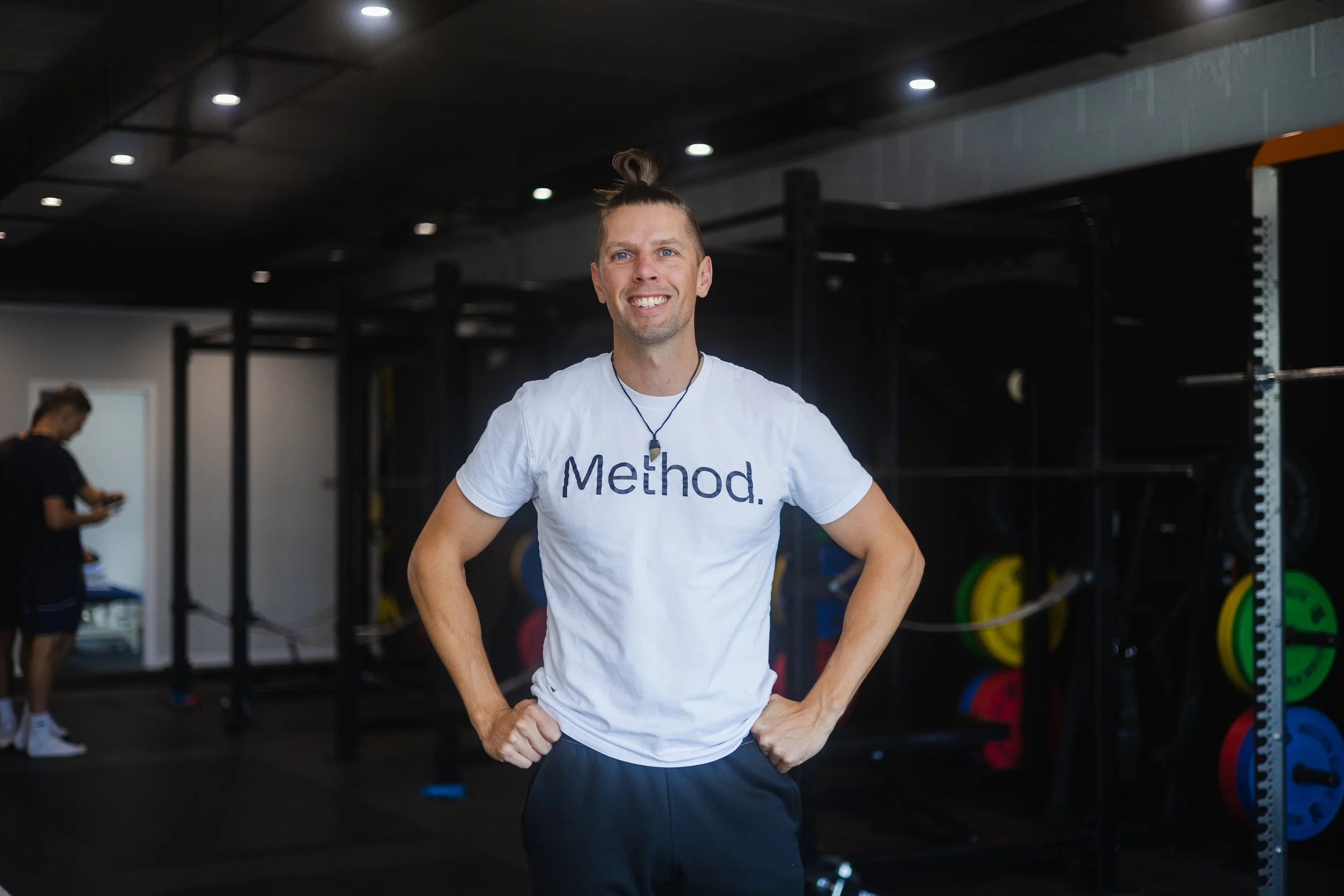 A man with long hair tied up in a bun, smiling, standing with hands on hips in a gym. He is wearing a white t-shirt with the word 'Method' printed on it and a necklace. The gym has weightlifting equipment, colorful weight plates, and another person in the background.