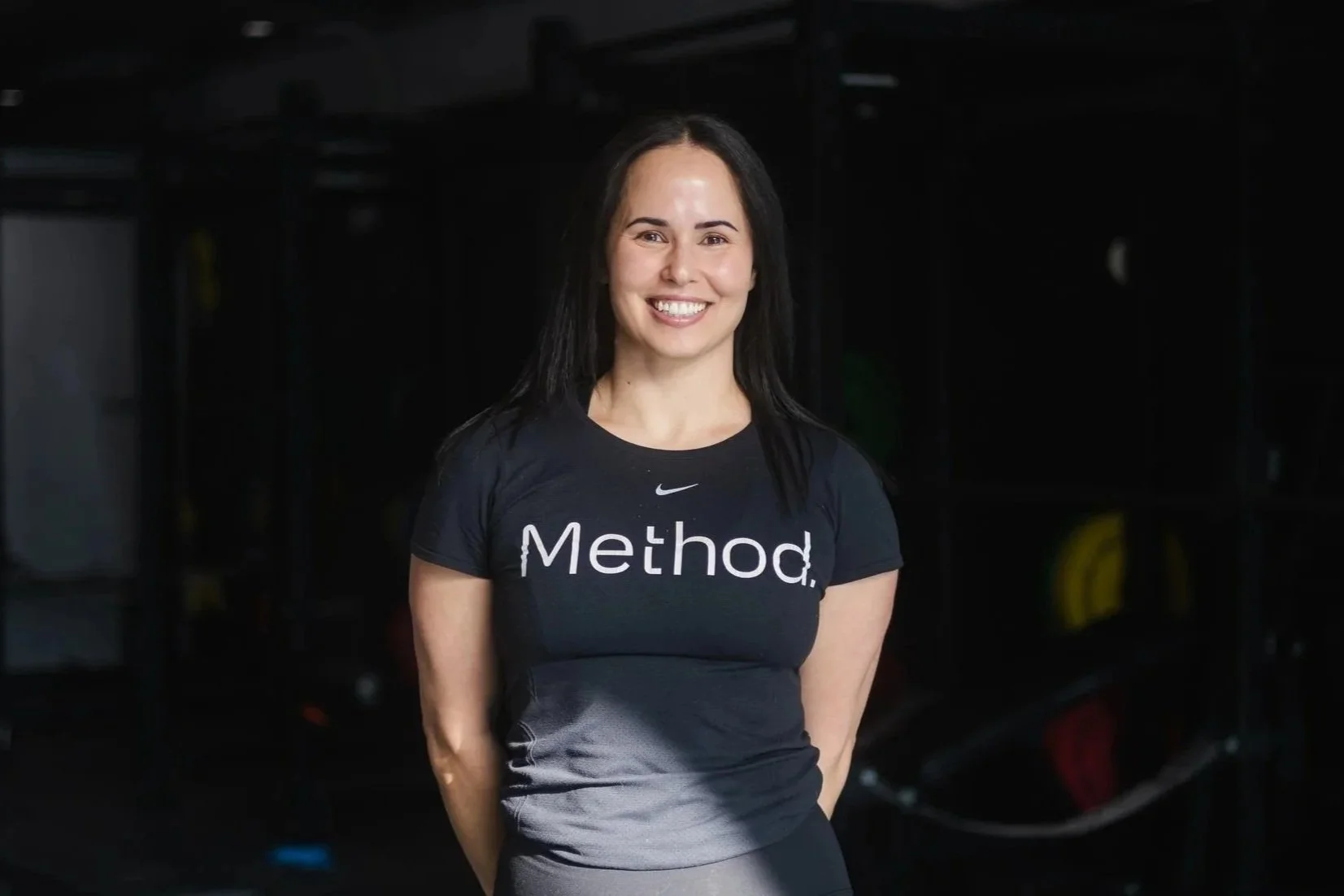 A woman with dark hair smiling at the camera, wearing a black T-shirt with the word "Method" printed on it in a gym setting.