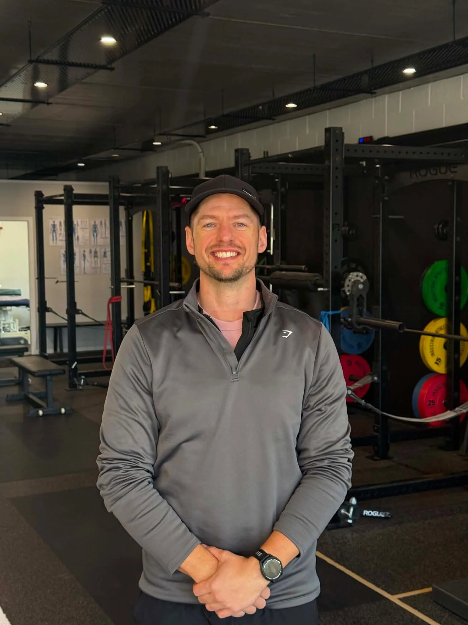 A man with light skin and a beard smiling in a gym, wearing a gray long-sleeve athletic shirt, black baseball cap, and watch, with fitness equipment including weights and a squat rack in the background.